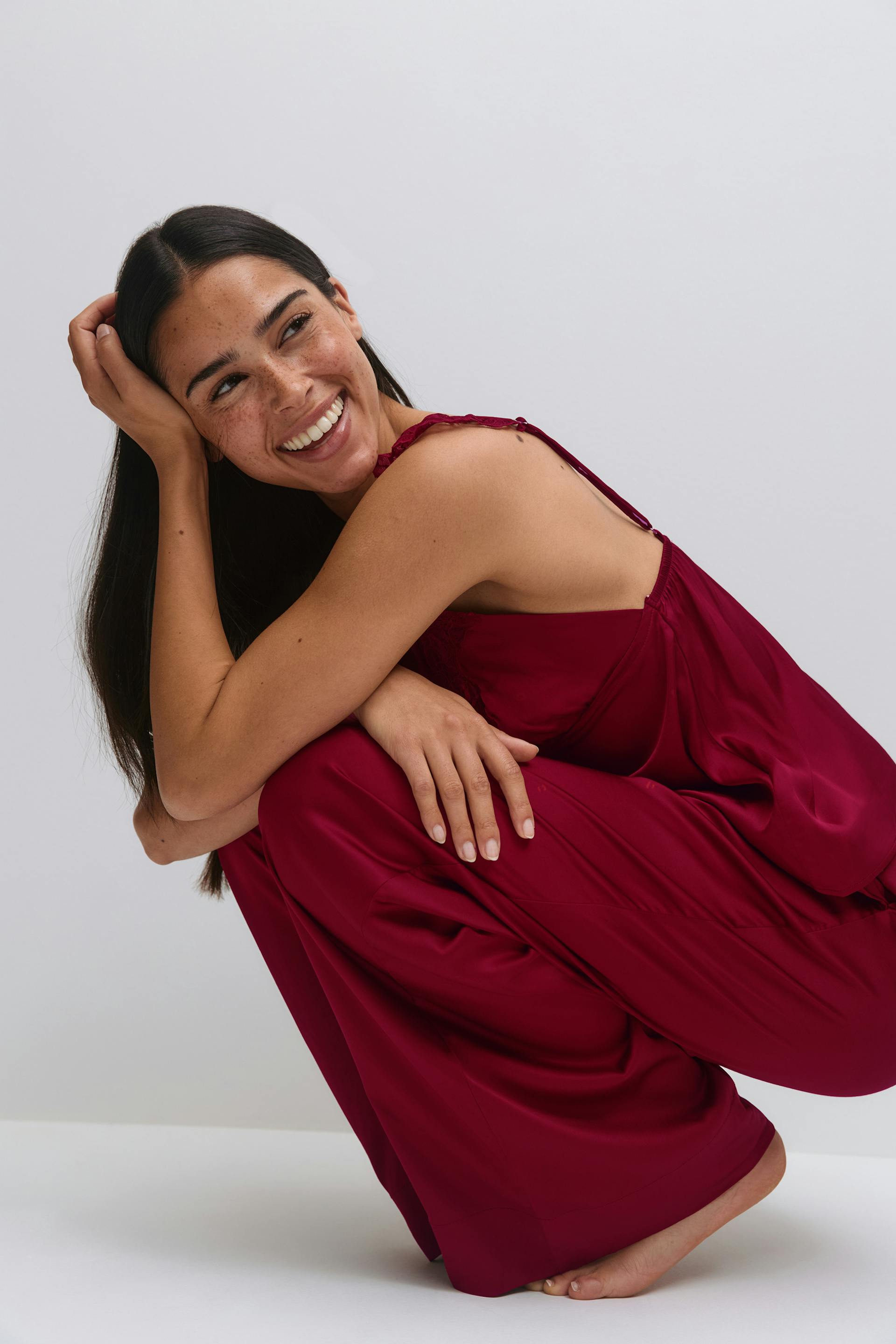 Deep burgundy satin camisole with lace‑trimmed adjustable spaghetti straps, paired with matching wide‑leg satin trousers; barefoot.