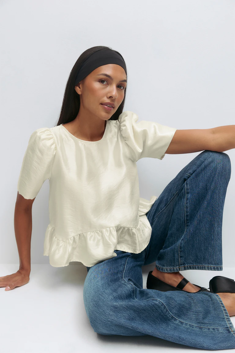 Kurzärmelige Bluse aus cremefarbenem Satin mit gepufften Schultern und gerüschtem Schößchensaum; mittelblaue Jeans mit weitem Bein; schwarzes breites Stirnband; schwarze Mary-Jane-Flats.