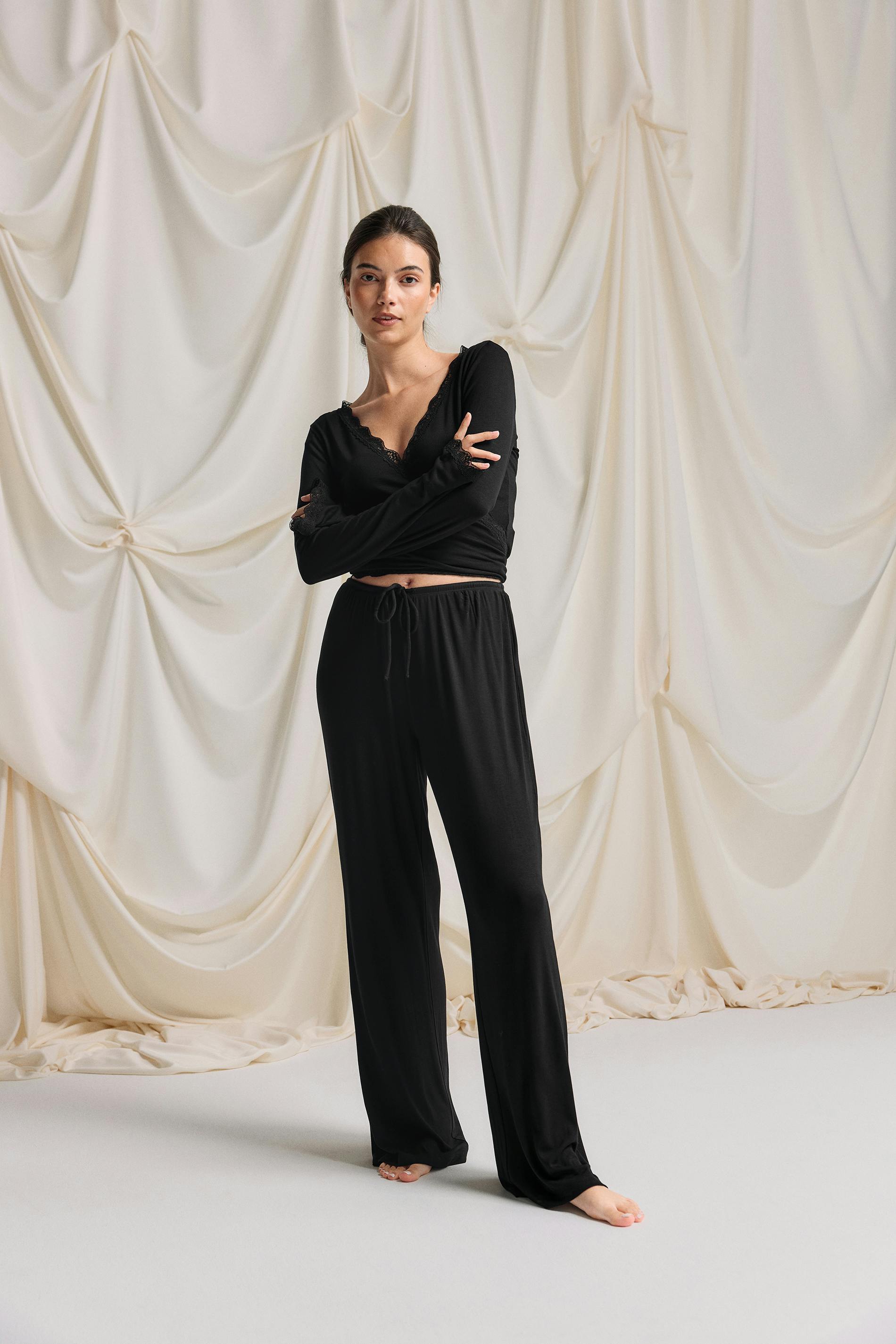 Svart langermet slå-om-topp med V-hals og mansjetter med blondekant, kombinert med matchende svart salongbukse med vidde ben og snøring; barbeint.