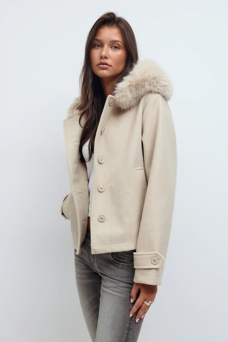 Beige uldjakke med knaplukning, pelskantet hætte, paspolerede lommer og manchetter med knaplukning, båret over en hvid top og sammen med grå slim-fit jeans.
