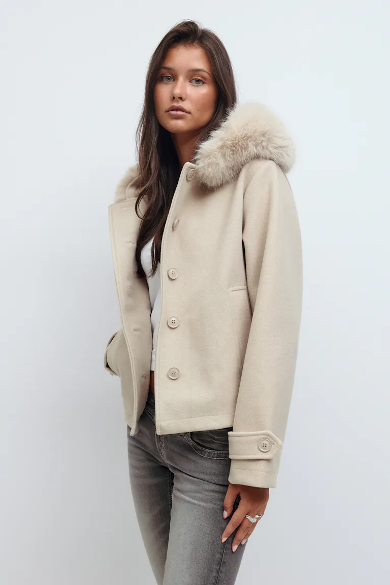 Valkoisen topin päälle puettava beige villatakki, jossa on turkisverhoiltu huppu, reunataskut ja napitettavat hihansuut. Yhdistettynä harmaisiin slim fit -farkkuihin.