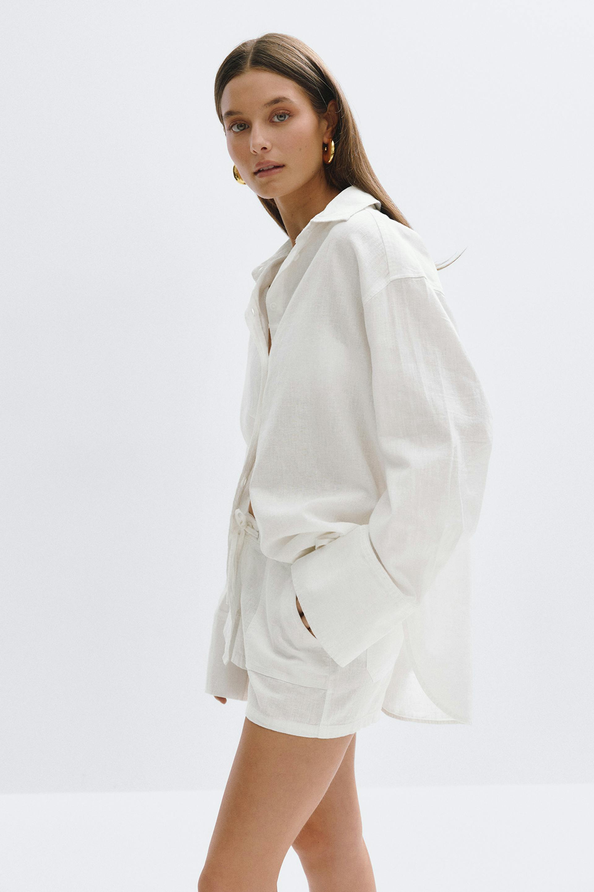 Chemise boutonnée en lin ivoire surdimensionnée à larges poignets, short en lin à cordon de serrage assorti et boucles d'oreilles en or.