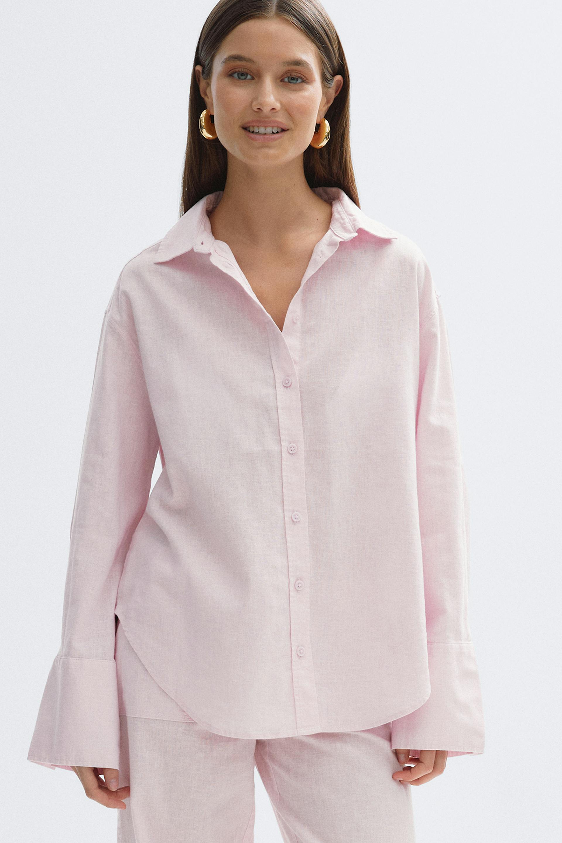 Chemise rose pâle à coupe décontractée et à poignets évasés, associée à un pantalon rose pâle assorti ; accessoirisée de grandes boucles d'oreilles dorées.