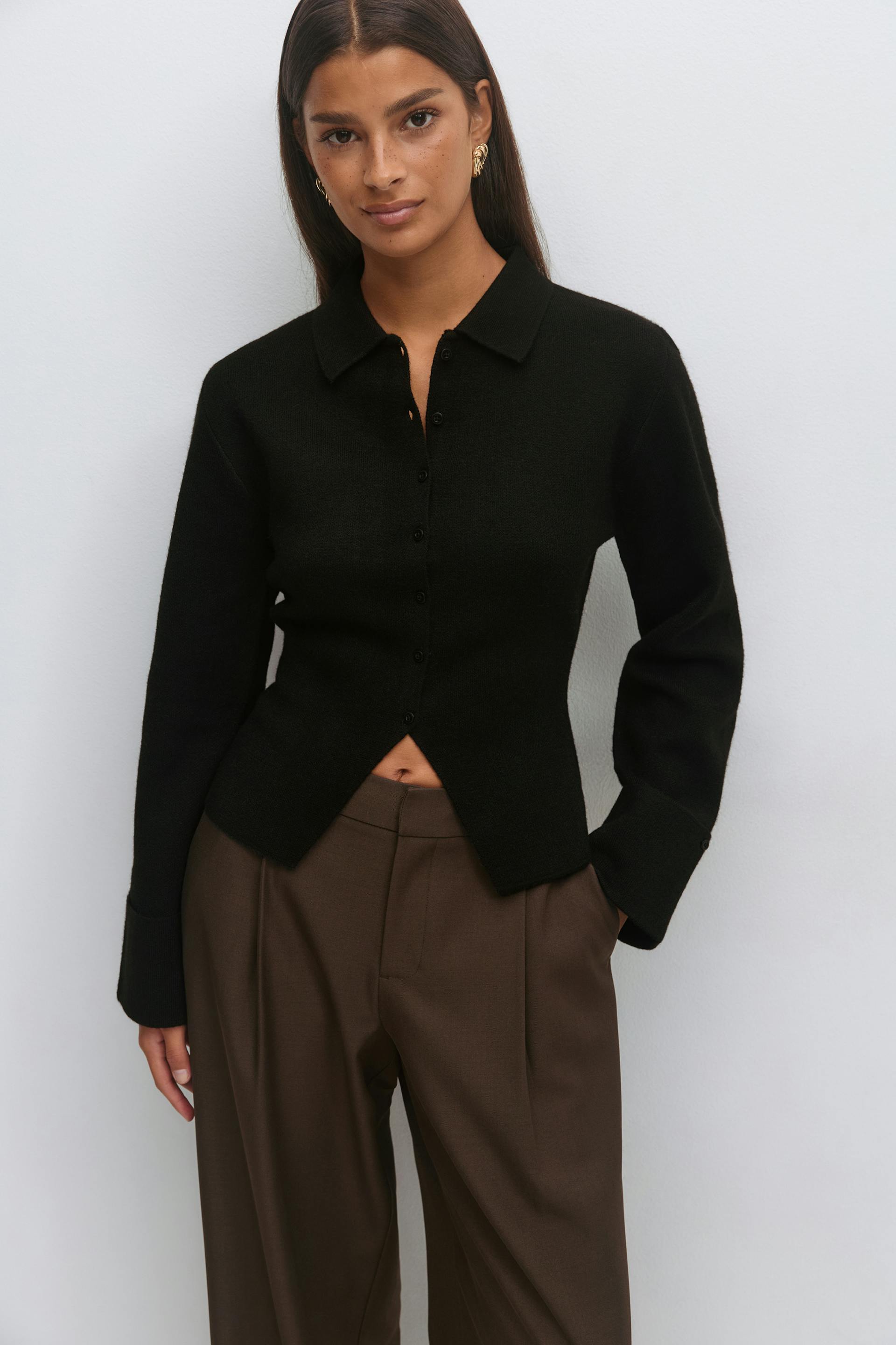 Black long-sleeve collared button-front knit cardigan with a triangular front hem opening, styled with high-waisted brown wide-leg trousers and gold sculptural earrings.