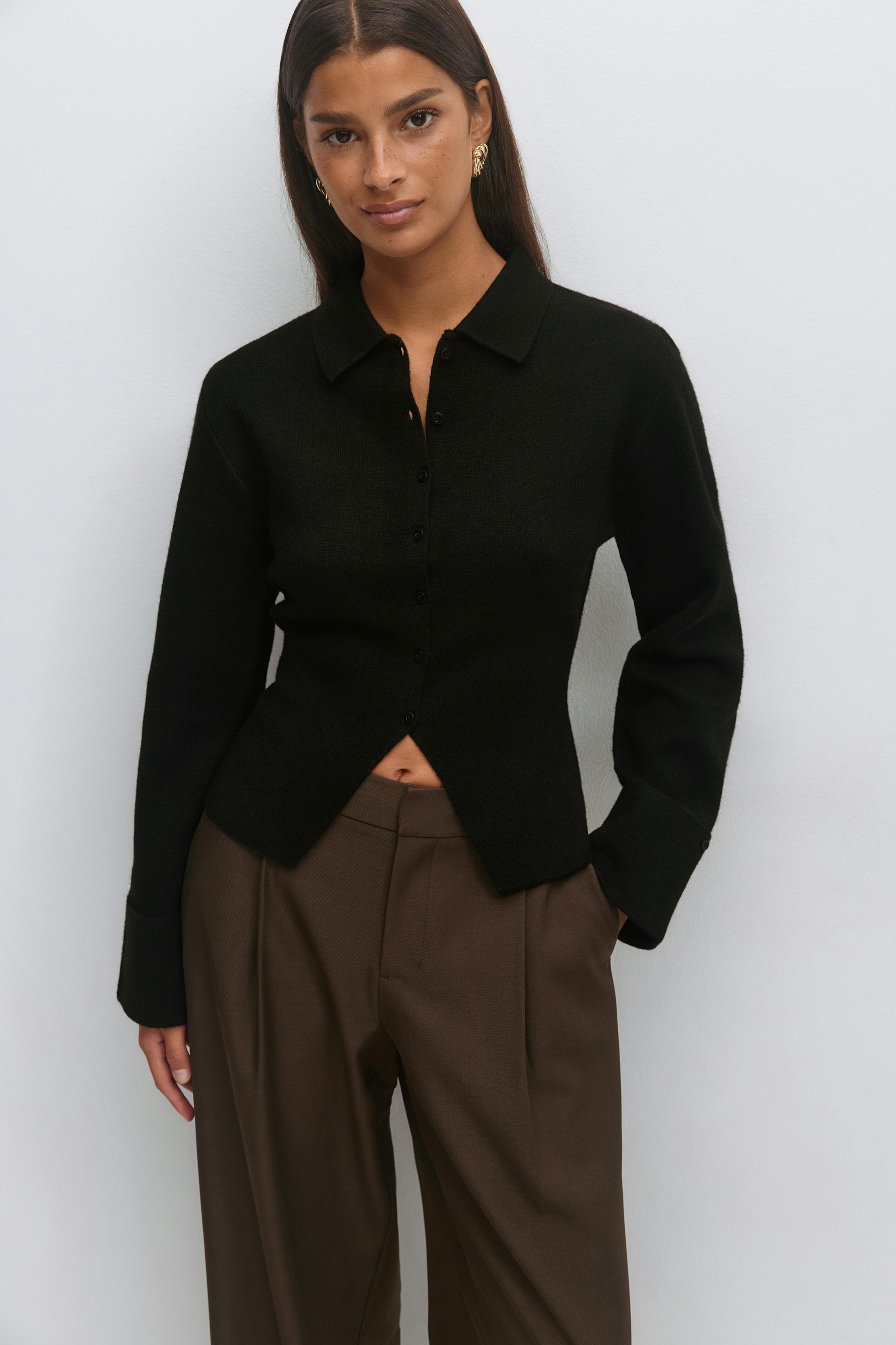 Black long-sleeve collared button-front knit cardigan with a triangular front hem opening, styled with high-waisted brown wide-leg trousers and gold sculptural earrings.
