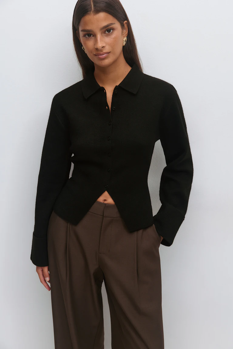 Cardigan noir à manches longues en maille boutonnée avec une ouverture triangulaire sur le devant, associé à un pantalon large marron à taille haute et à des boucles d'oreilles sculpturales dorées.