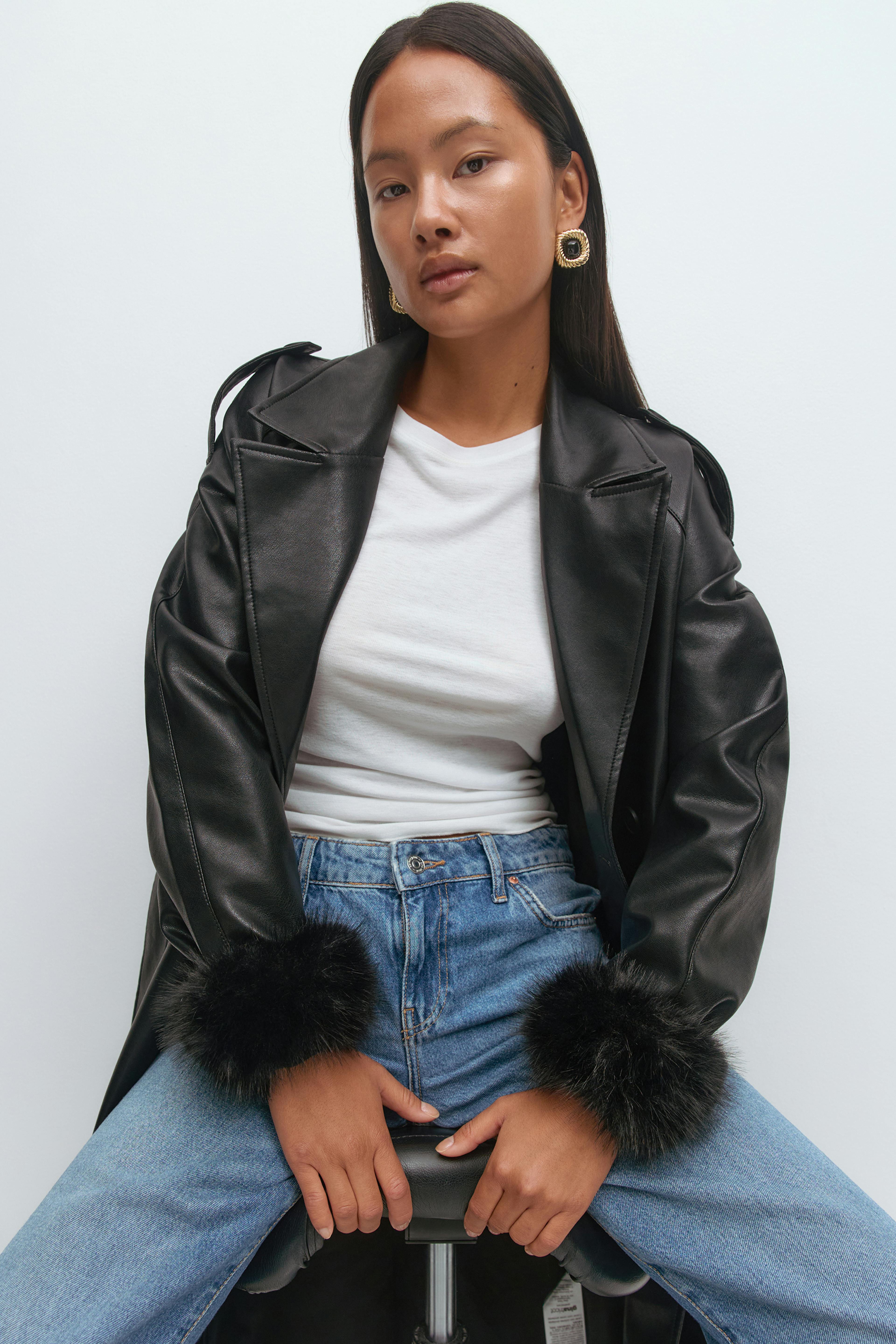 Black leather-effect trench coat with black fur cuffs, white crew-neck T-shirt, light blue high-waisted jeans, and gold statement hoop earrings.