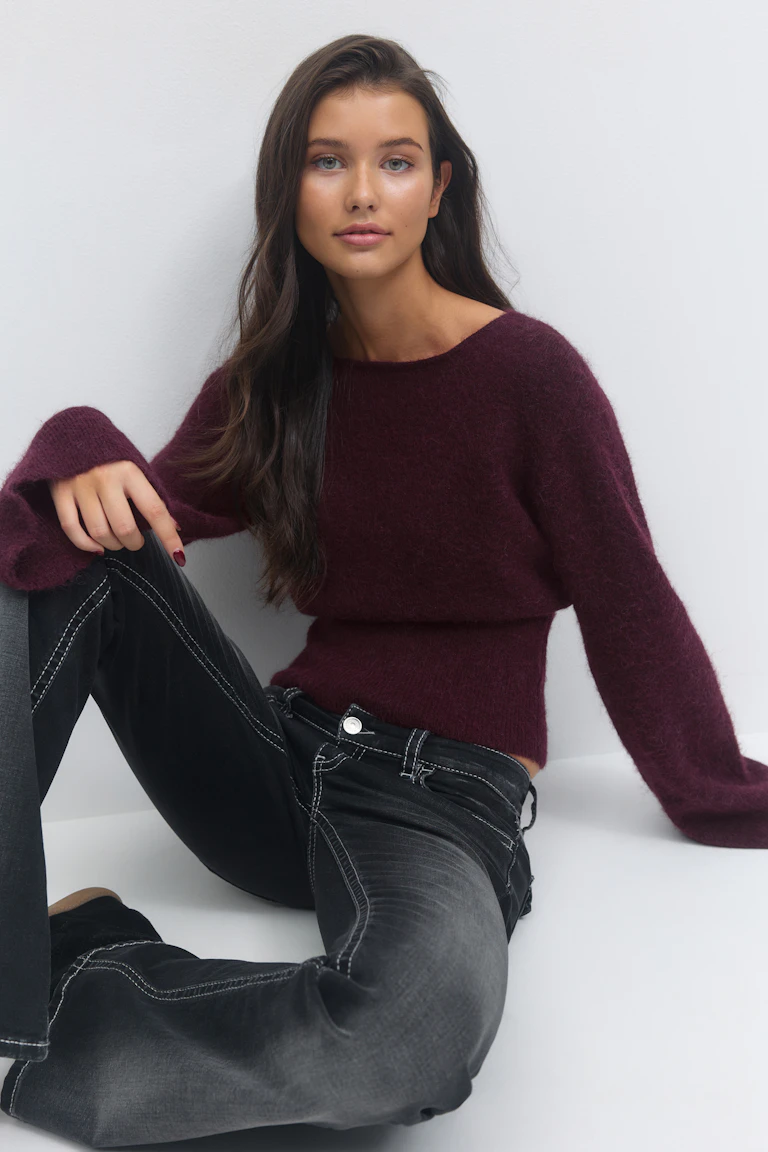 Een diep bordeauxrode pluizige trui met lange mouwen en een boothals gecombineerd met een vervaagde zwarte jeans met witte contraststiksels.