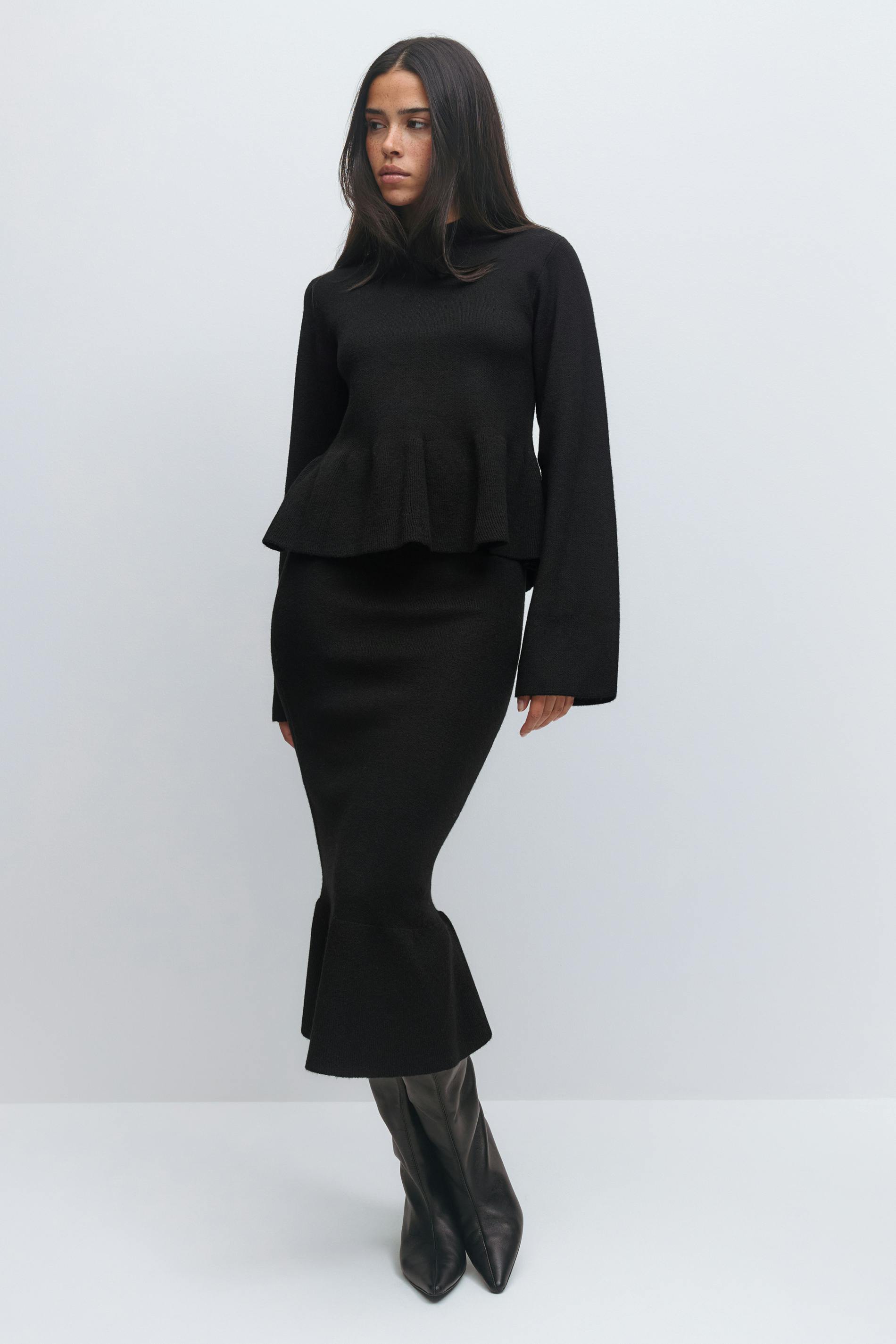 Femme portant un pull peplum noir à manches longues et une jupe midi noire ajustée à l'ourlet évasé, associée à des bottes en cuir noires à bout pointu.