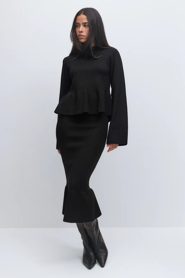 Kvinde iført en sort strikket peplum-sweater med lange ærmer og en tætsiddende sort midi-nederdel med udslået kant, parret med sorte spidse, knæhøje læderstøvler.