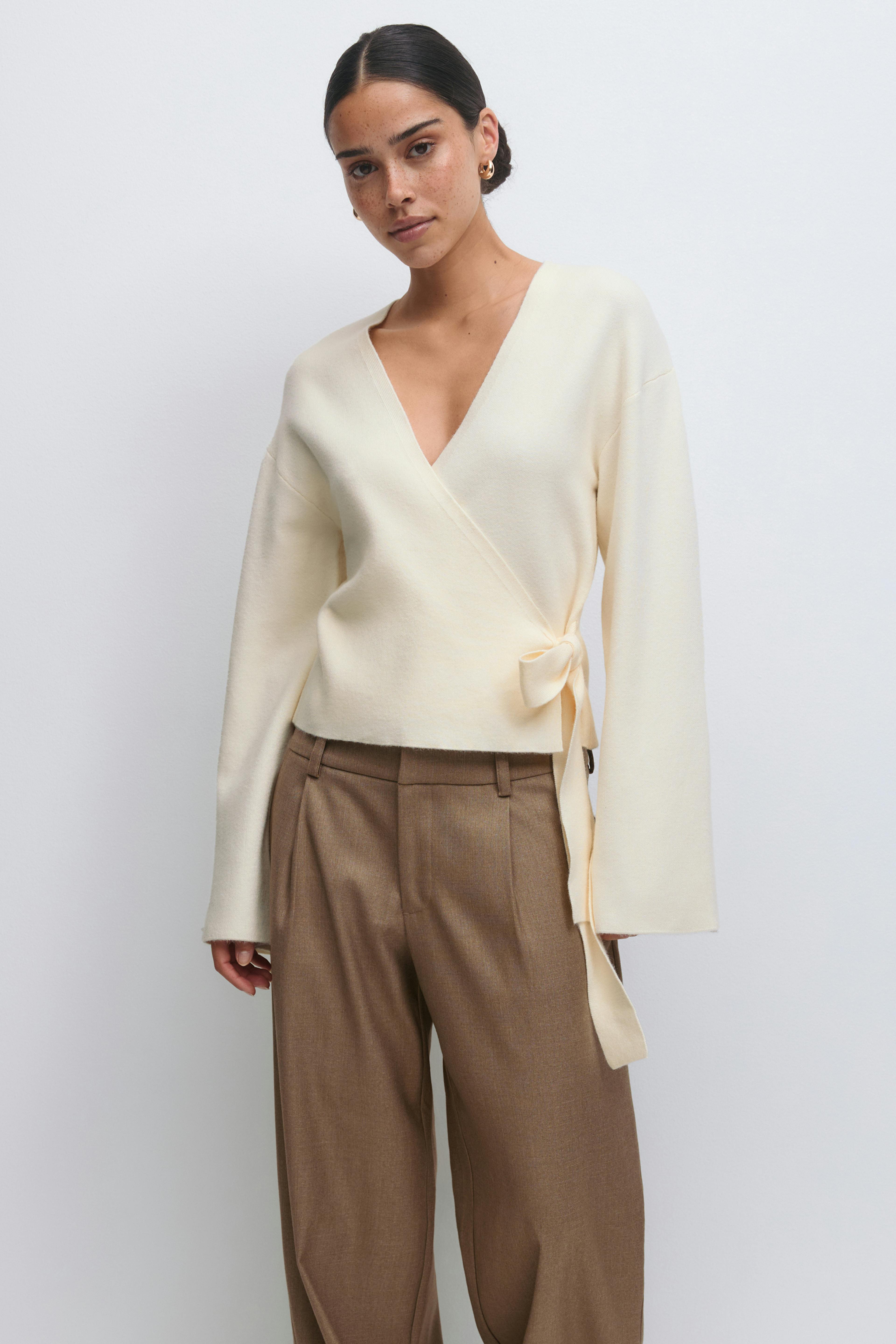 Kermanvärinen V-kauluksinen pusero, jossa on pitkät hihat ja sivussa solmittava ruskea ruskeat leveälahkeiset laskostetut korkeavyötäröiset housut.