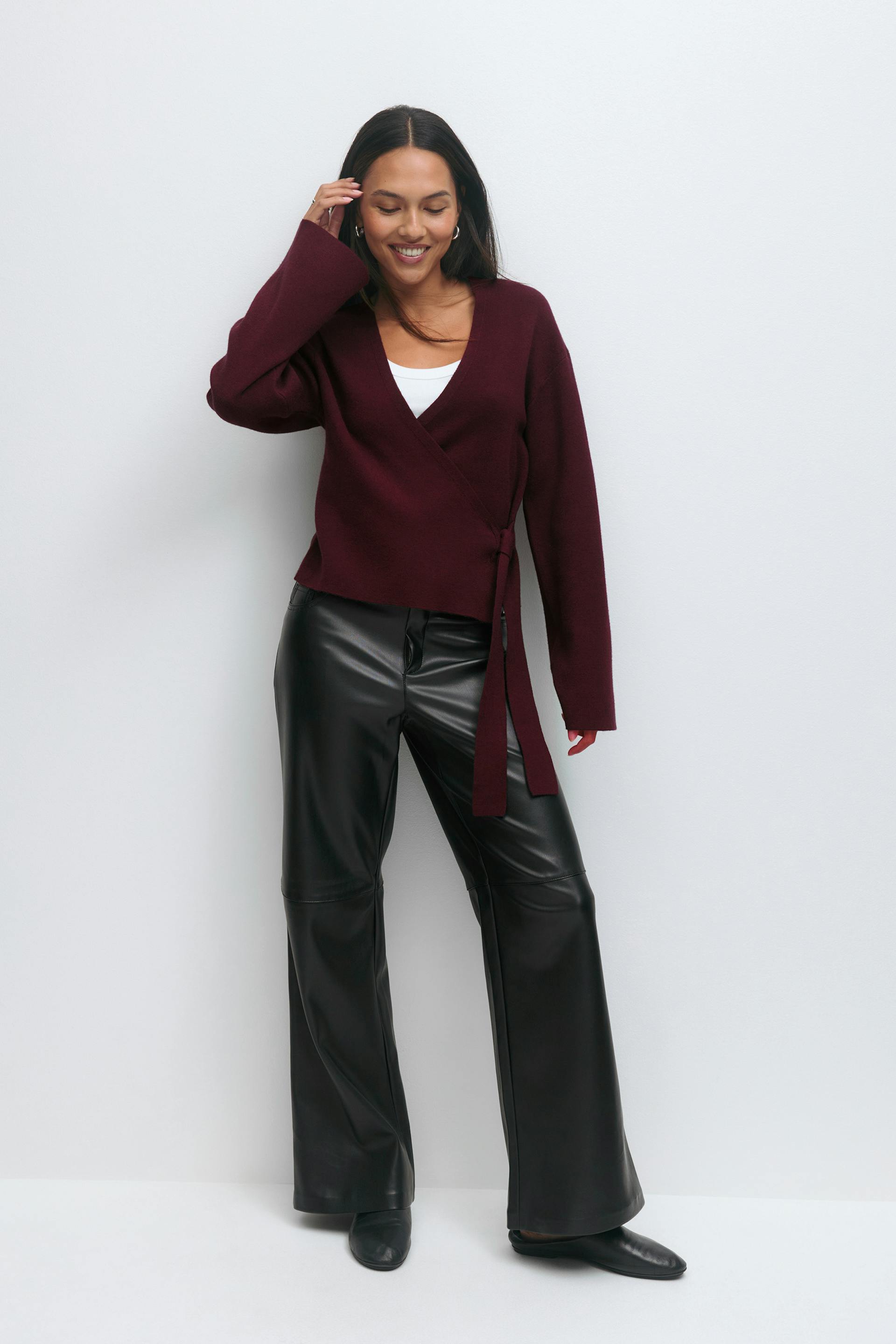 Het model draagt een bordeauxrode wikkeltrui met stropdas aan de zijkant over een witte tank met ronde hals, een zwarte broek met wijde pijpen van imitatieleer en zwarte enkellaarsjes.