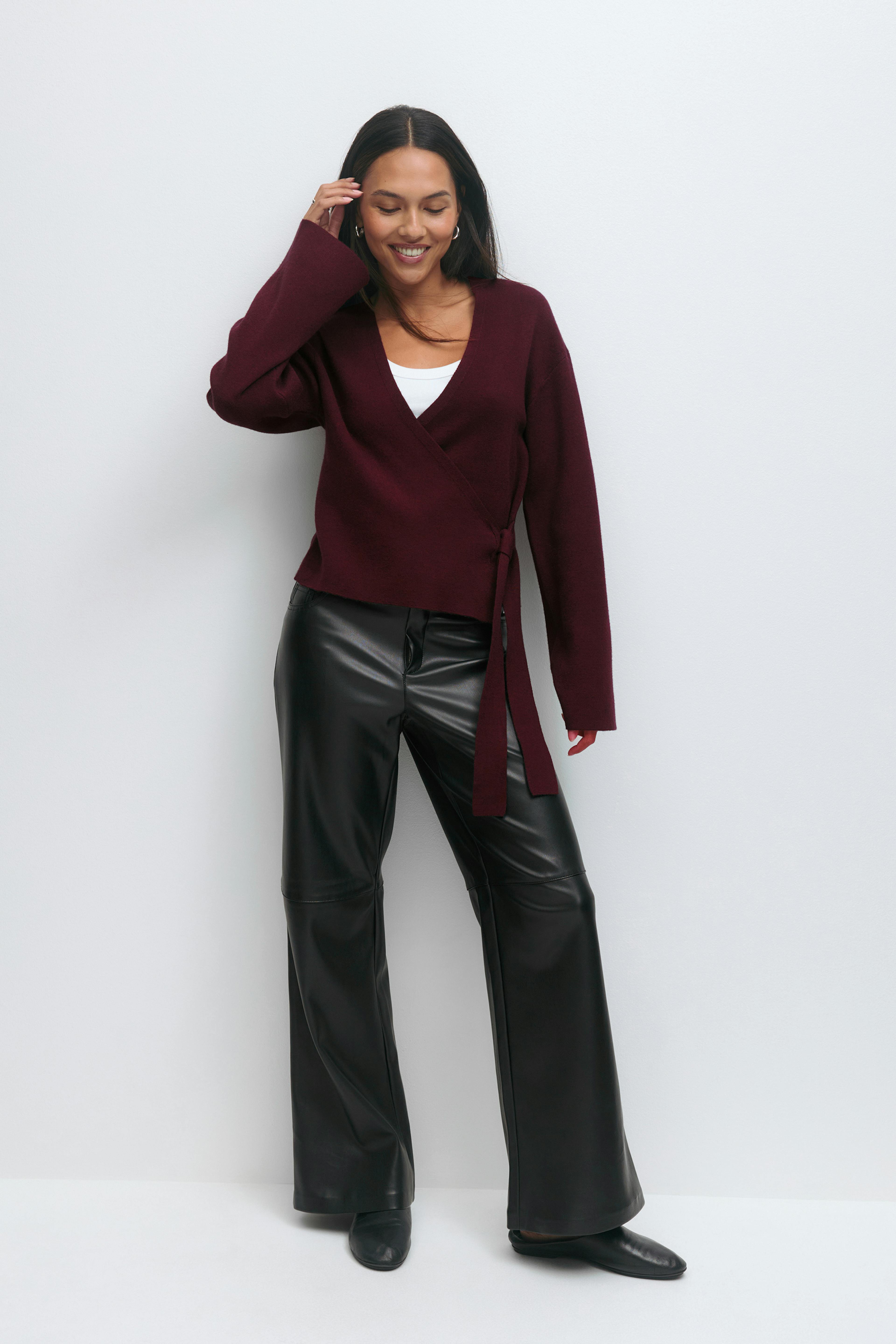 Modellen bär en vinröd omlottröja med sidoslips över ett vitt linne med rund halsringning, svarta vida byxor i konstläder och svarta ankelstövlar.