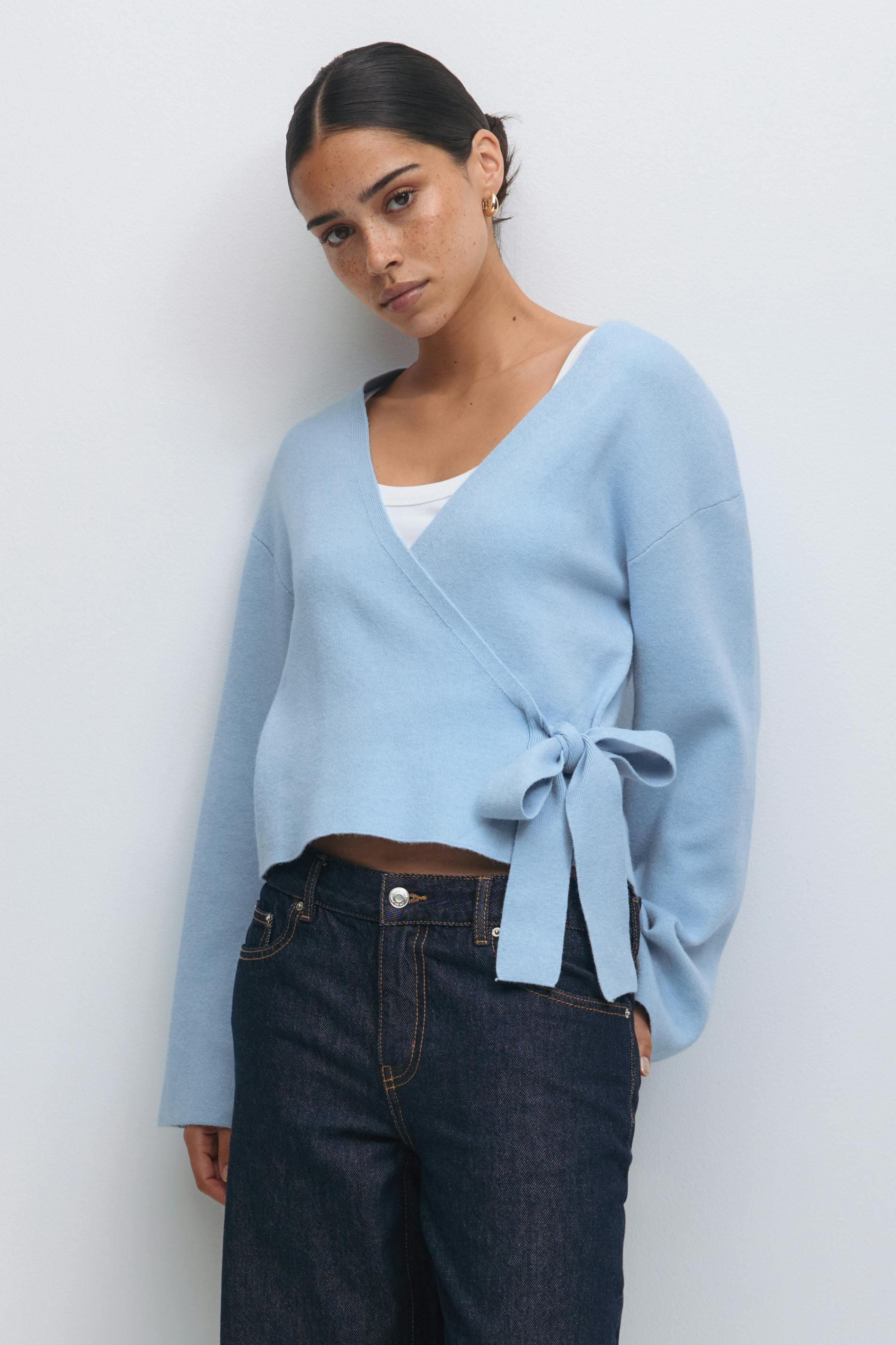 Lichtblauwe overslagtrui met V-hals, lange mouwen en een strikje aan de zijkant, in laagjes over een wit hemdje, gecombineerd met een indigo-donkere jeans met hoge taille en kleine gouden oorbellen.