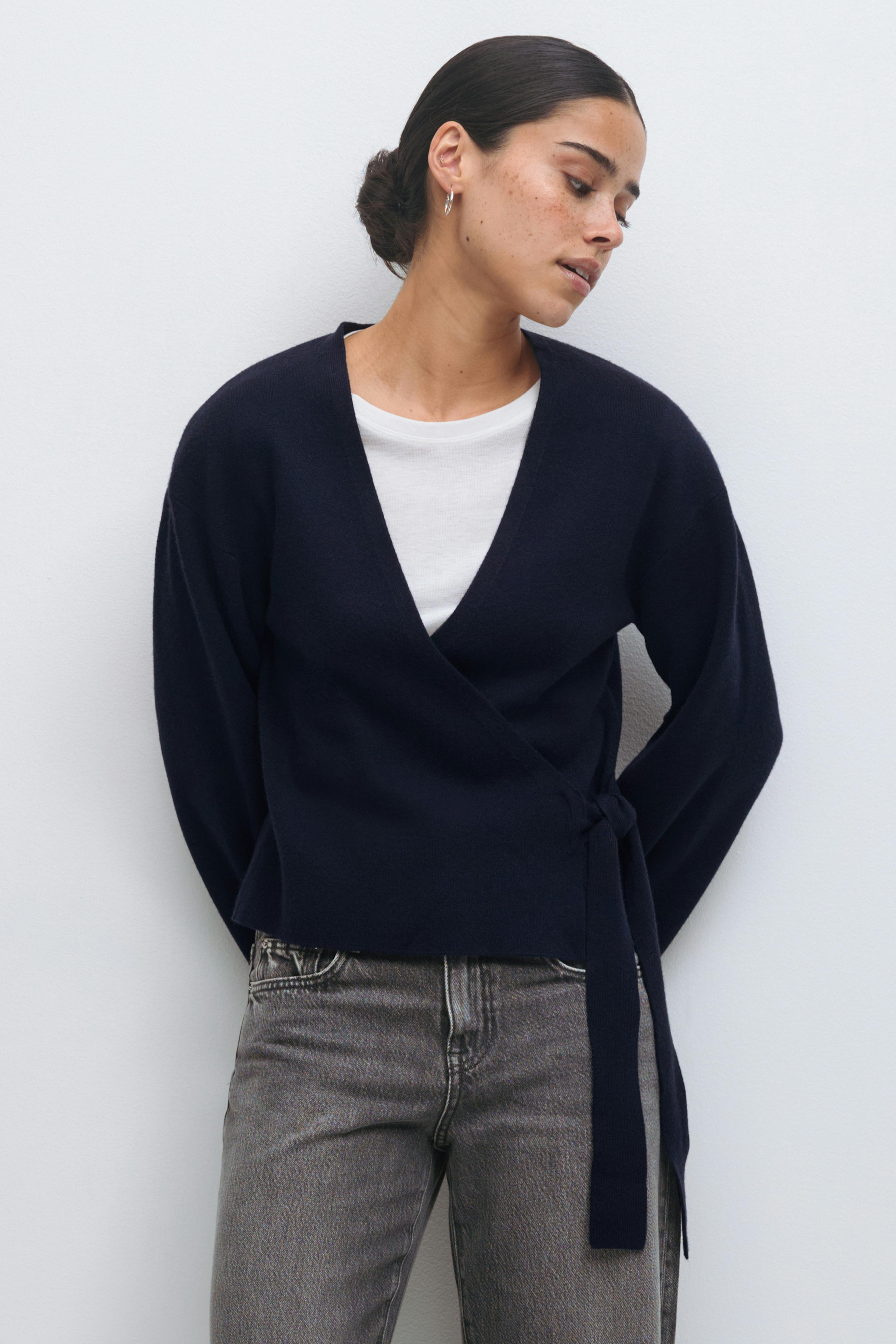 Marineblaue Wickeljacke mit seitlicher Bindung über einem weißen T-Shirt mit Rundhalsausschnitt, kombiniert mit verblichenen grauen High-Waist-Jeans und kleinen silbernen Ohrringen.