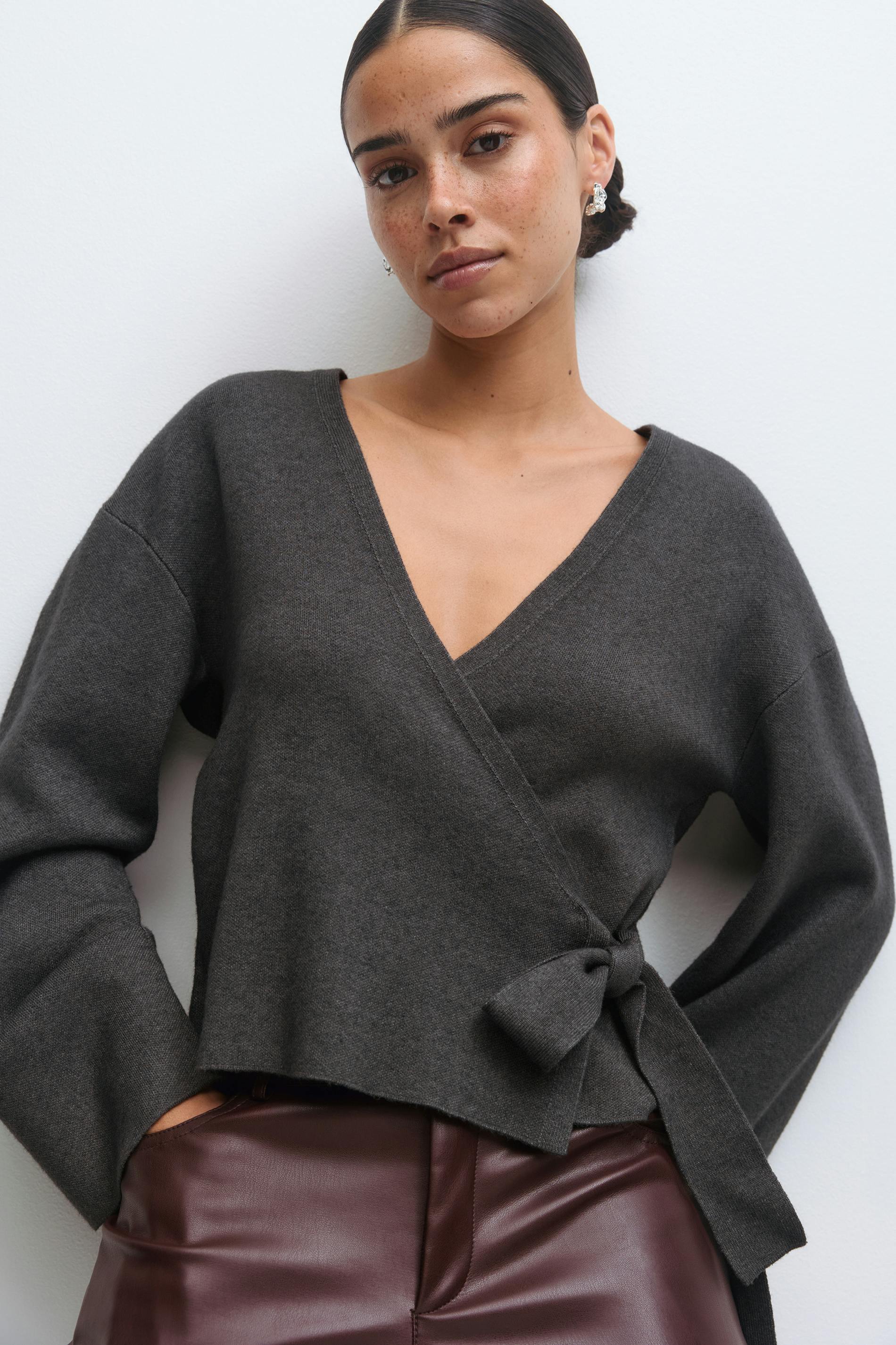 Charcoal wrap V-neck sweater with long sleeves and a side tie, paired with high-waisted burgundy leather trousers.