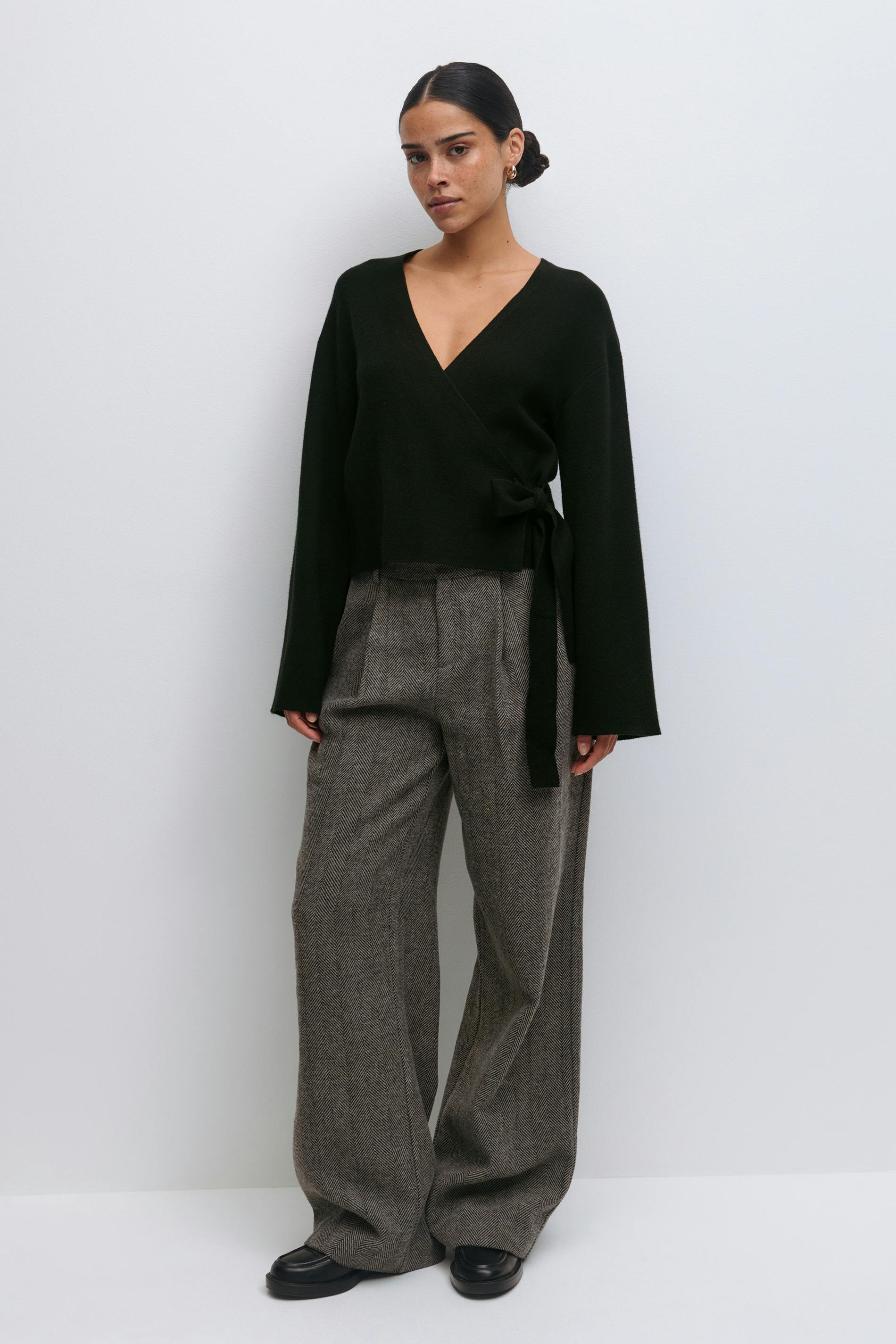 Schwarzer Wickelpullover mit V-Ausschnitt, langen Ärmeln und seitlicher Bindung, kombiniert mit einer braunen Fischgrätenhose mit hoher Taille und schwarzen Leder-Slippern.