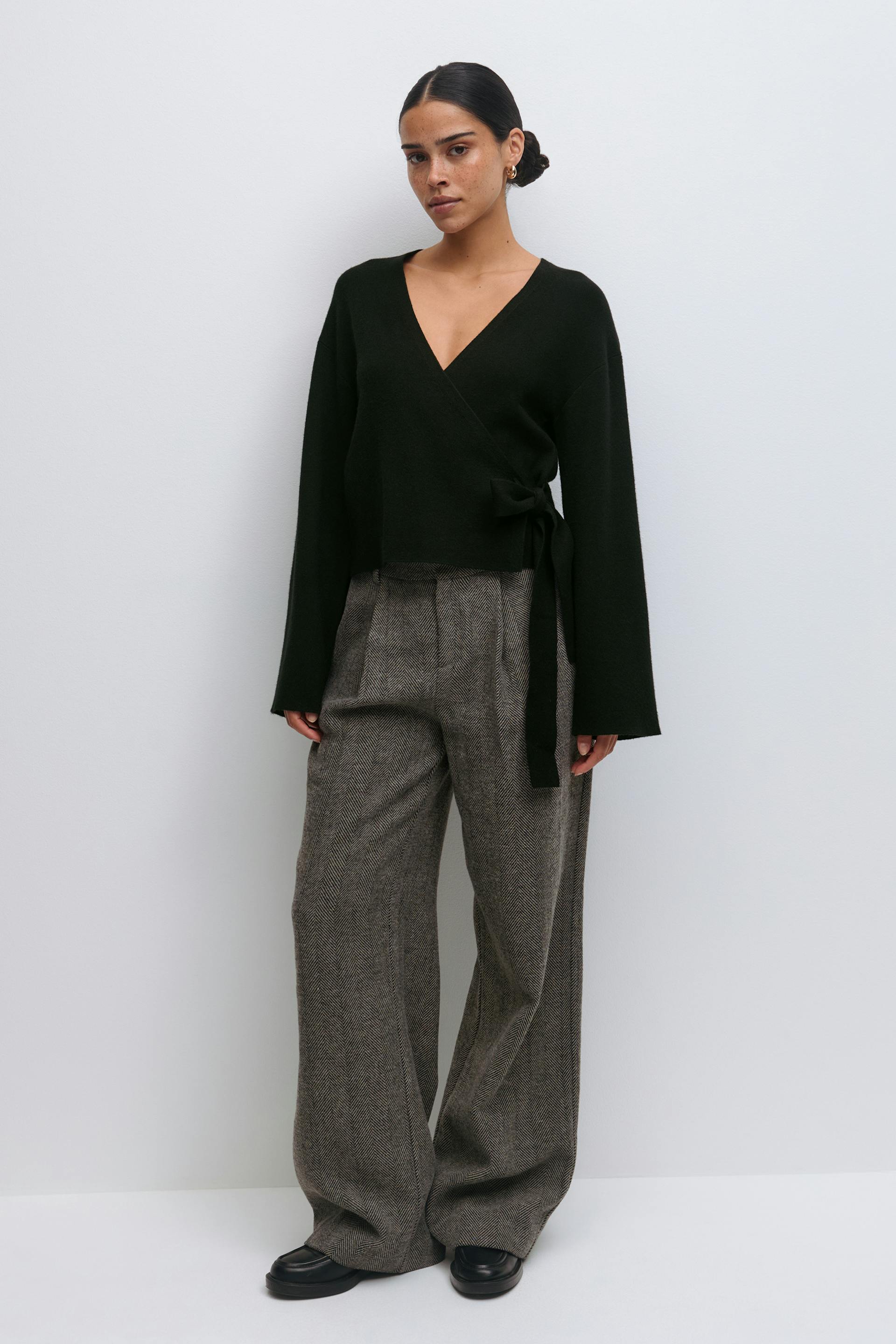 Zwarte overslagtrui met V-hals, lange mouwen en een striksluiting aan de zijkant, gecombineerd met een bruine visgraatbroek met hoge taille en zwarte leren loafers.