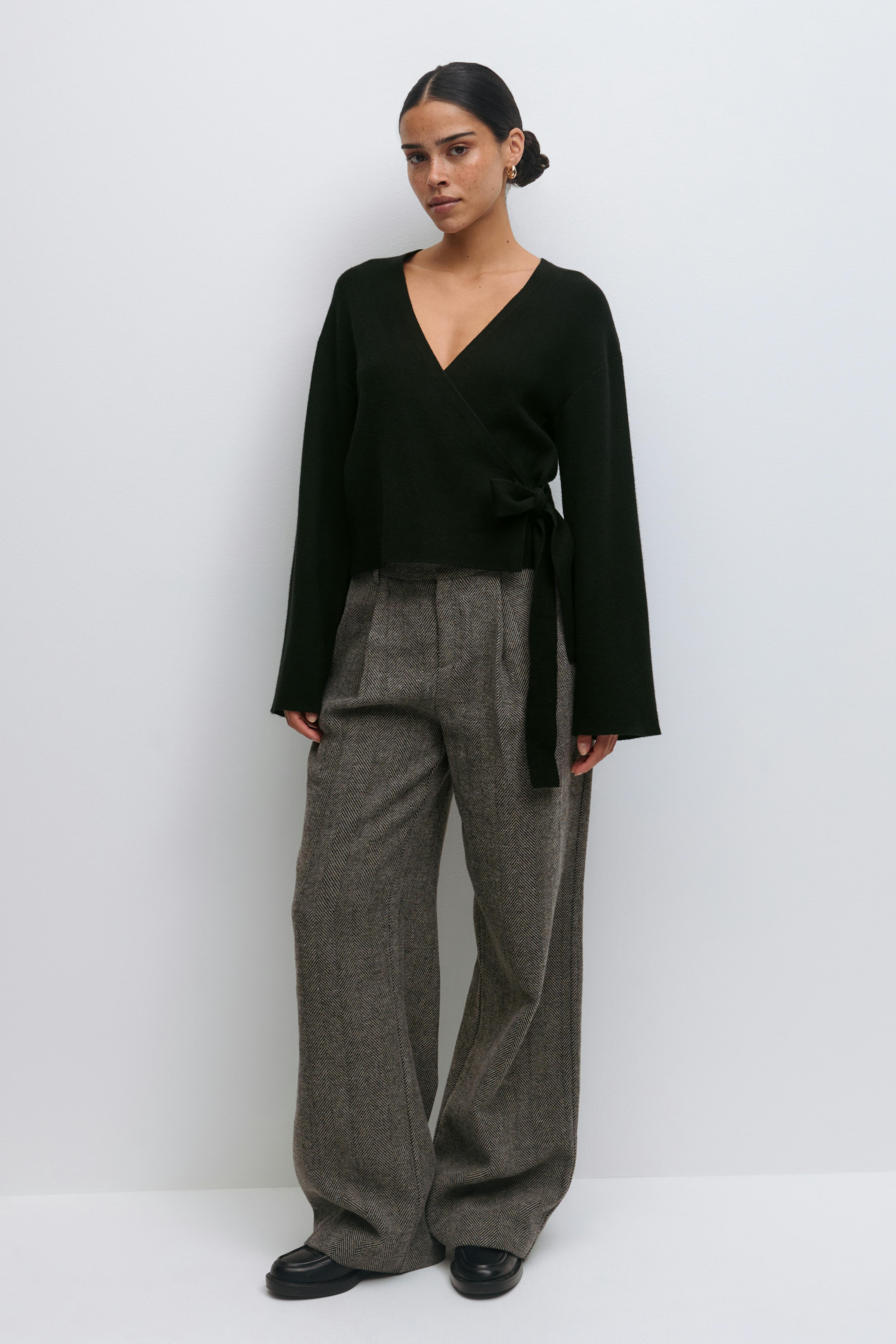Schwarzer Wickelpullover mit V-Ausschnitt, langen Ärmeln und seitlicher Bindung, kombiniert mit einer braunen Fischgrätenhose mit hoher Taille und schwarzen Leder-Slippern.