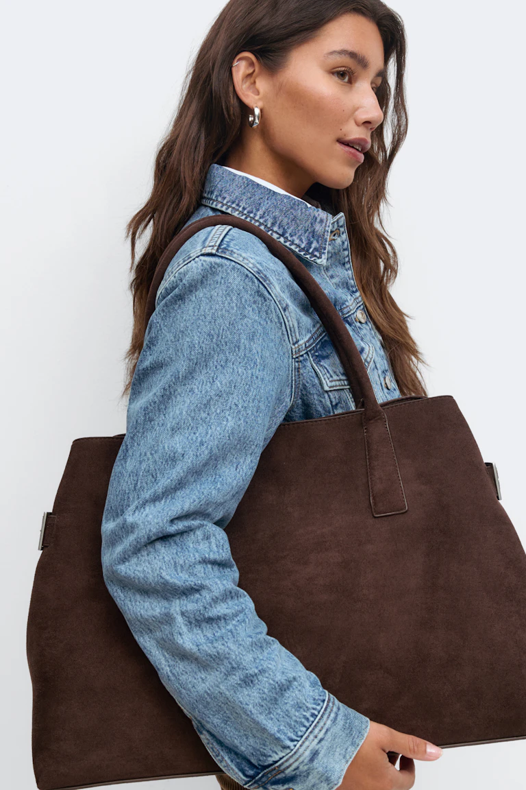 Veste en jean bleu clair sur un haut blanc, boucles d'oreilles argentées et grand sac fourre-tout en daim marron avec bandoulières.