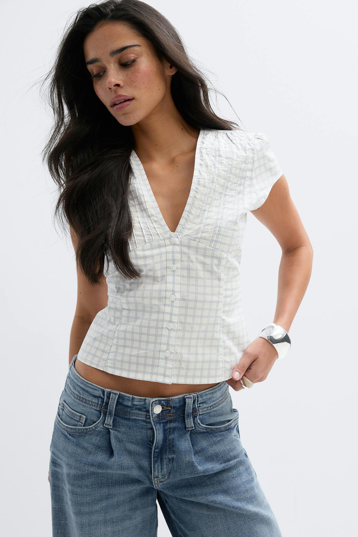 Vit kortärmad blus med V-ringad knappslå och ljusblått rutmönster, kombinerad med jeans med avslappnad passform i mellanblå färg; kompletterad med ett silverfärgat armband och ring.