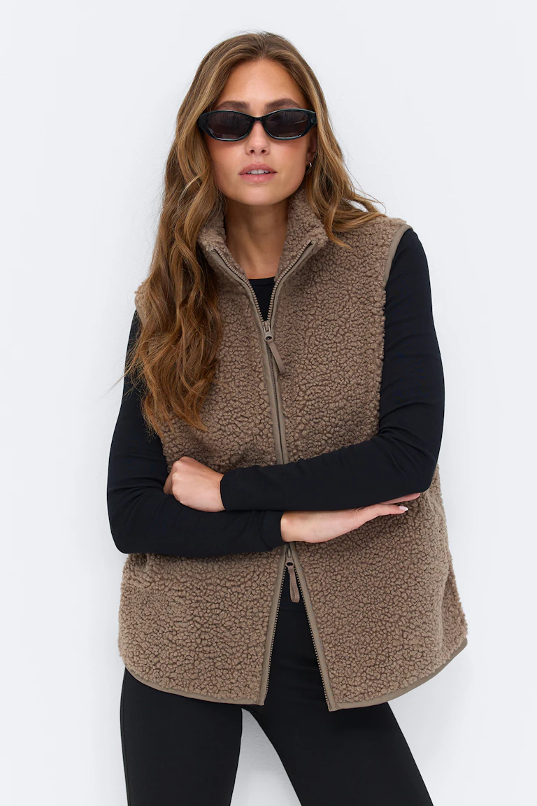 Brown sherpa zip-front sleeveless vest worn over a black long-sleeve top, paired with black leggings and slim black cat-eye sunglasses.