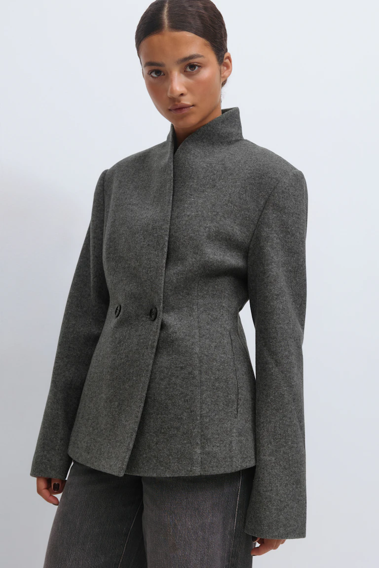 Anthrazitgraue, maßgeschneiderte Wolljacke mit hohem Stehkragen, verdeckter Knopfleiste mit zwei Knöpfen in der Taille und langen Ärmeln, kombiniert mit dunkelgrauen Jeans.