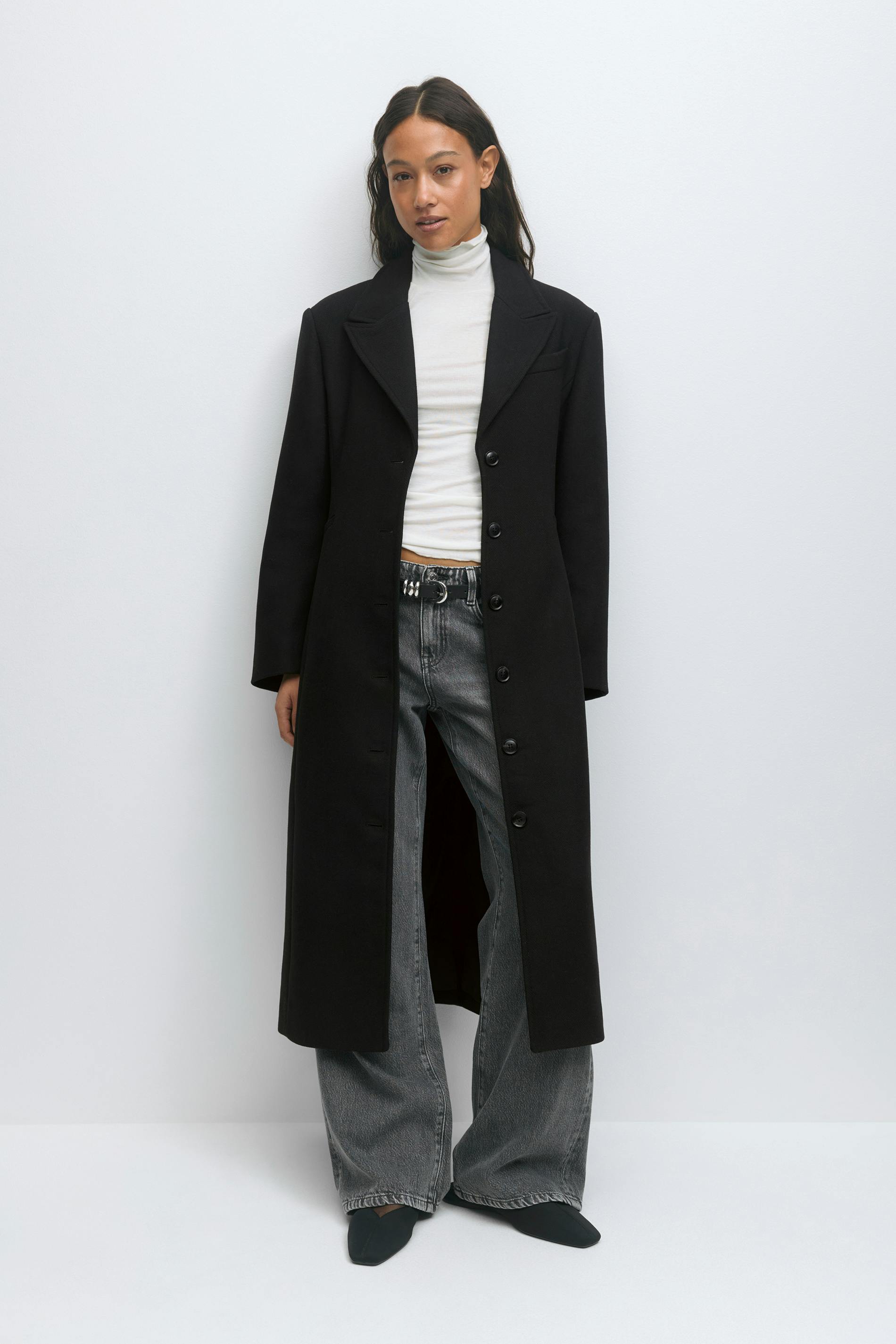 Pitkä musta yksirivinen nappitakki, jota käytetään valkoisen poolokauluksen päällä, yhdistettynä haalistuneisiin harmaisiin leveälahkeisiin farkkuihin, mustaan nastavyöhön ja mustiin teräväkärkisiin kenkiin.