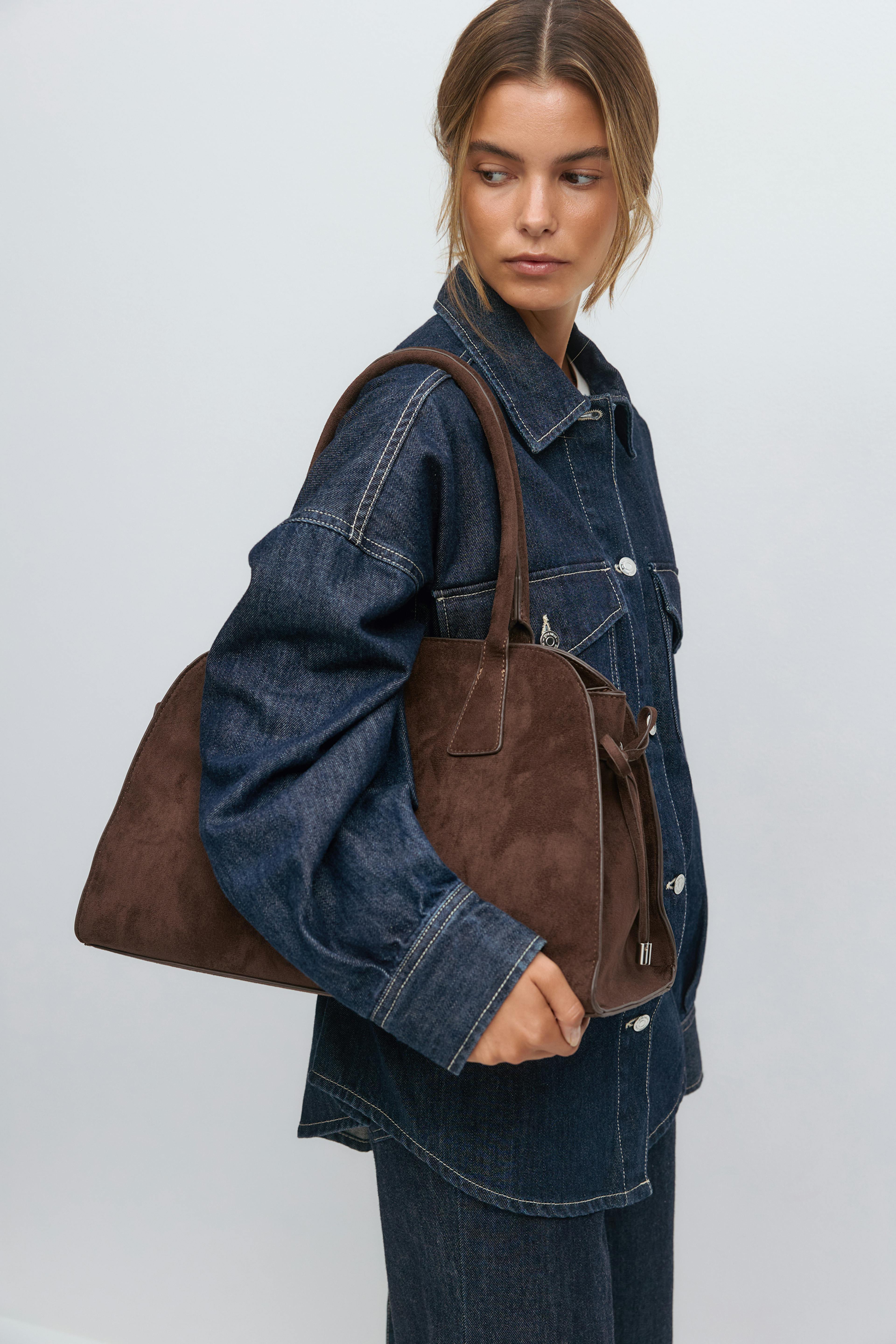 Mørk indigo denimjakke med hvide kontrastsyninger, matchende denimbukser og en struktureret tote-taske i brunt ruskind med dobbelt håndtag.