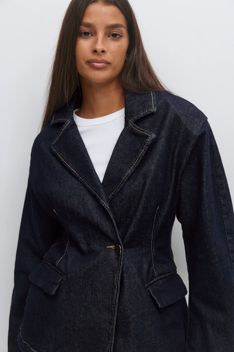 Blazer en denim indigo foncé avec revers cranté, surpiqûres contrastées, fermeture à bouton unique et poches à rabat, superposé à un T-shirt blanc à col ras du cou.