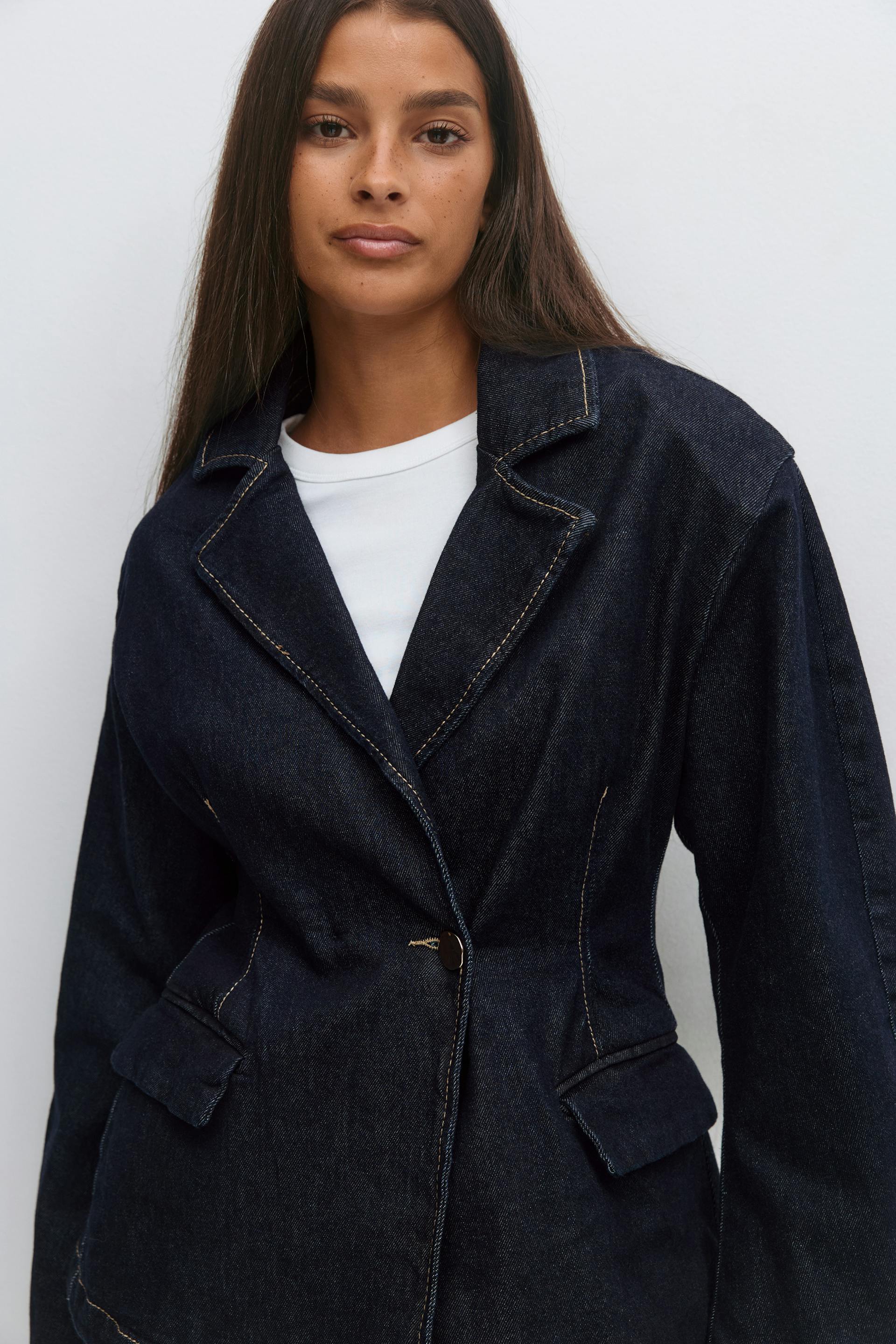 Dark indigo denim blazer with notched lapel, contrast stitching, single-button closure and flap pockets, layered over a white crew-neck T‑shirt.