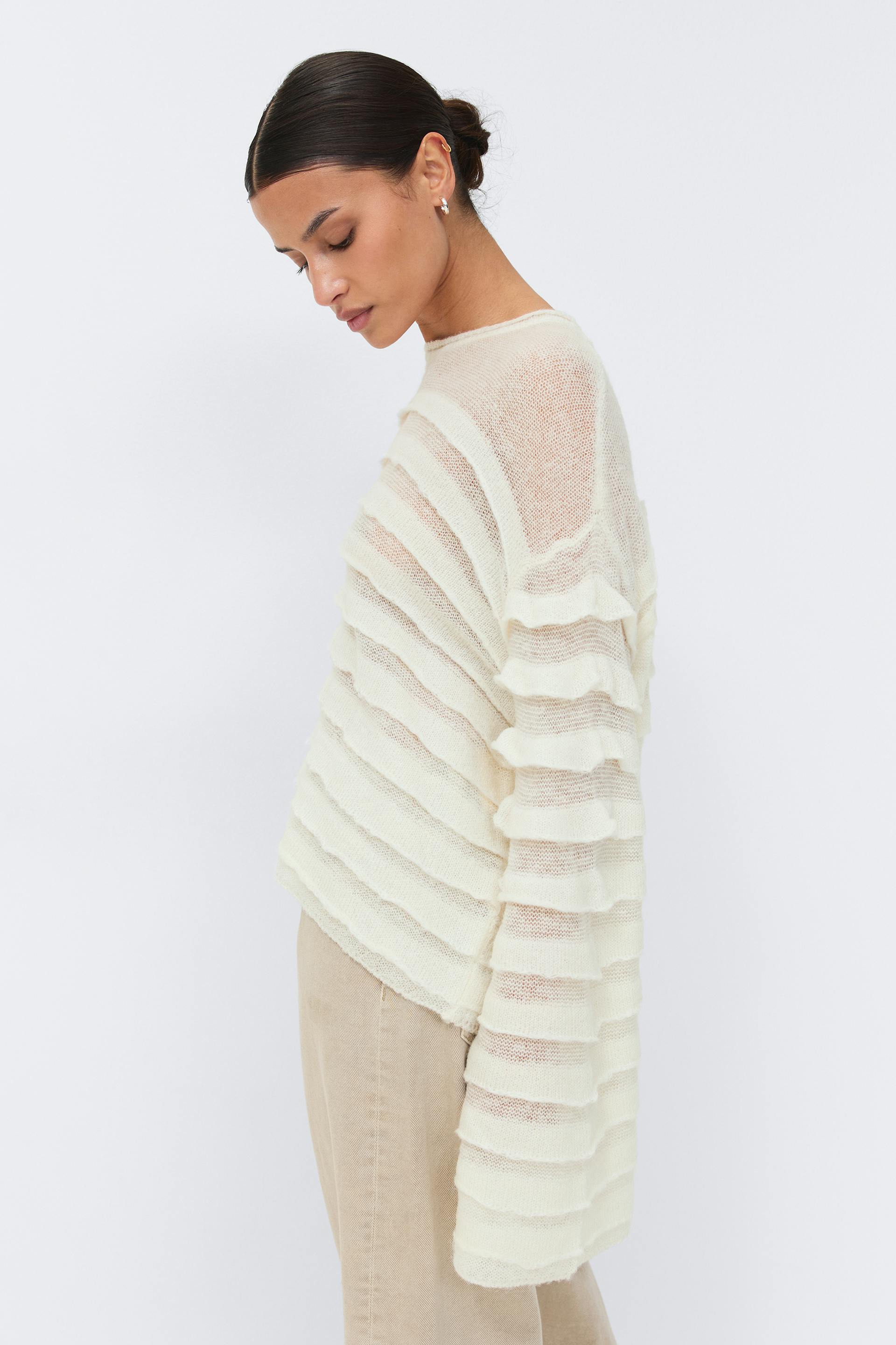 Norsunluunvärinen, puoliksi läpikuultava neulepusero, jossa on vaakasuuntaiset röyhelöraidat ja pitkät hihat, yhdistettynä beigeen korkeavyötäröisiin housuihin.