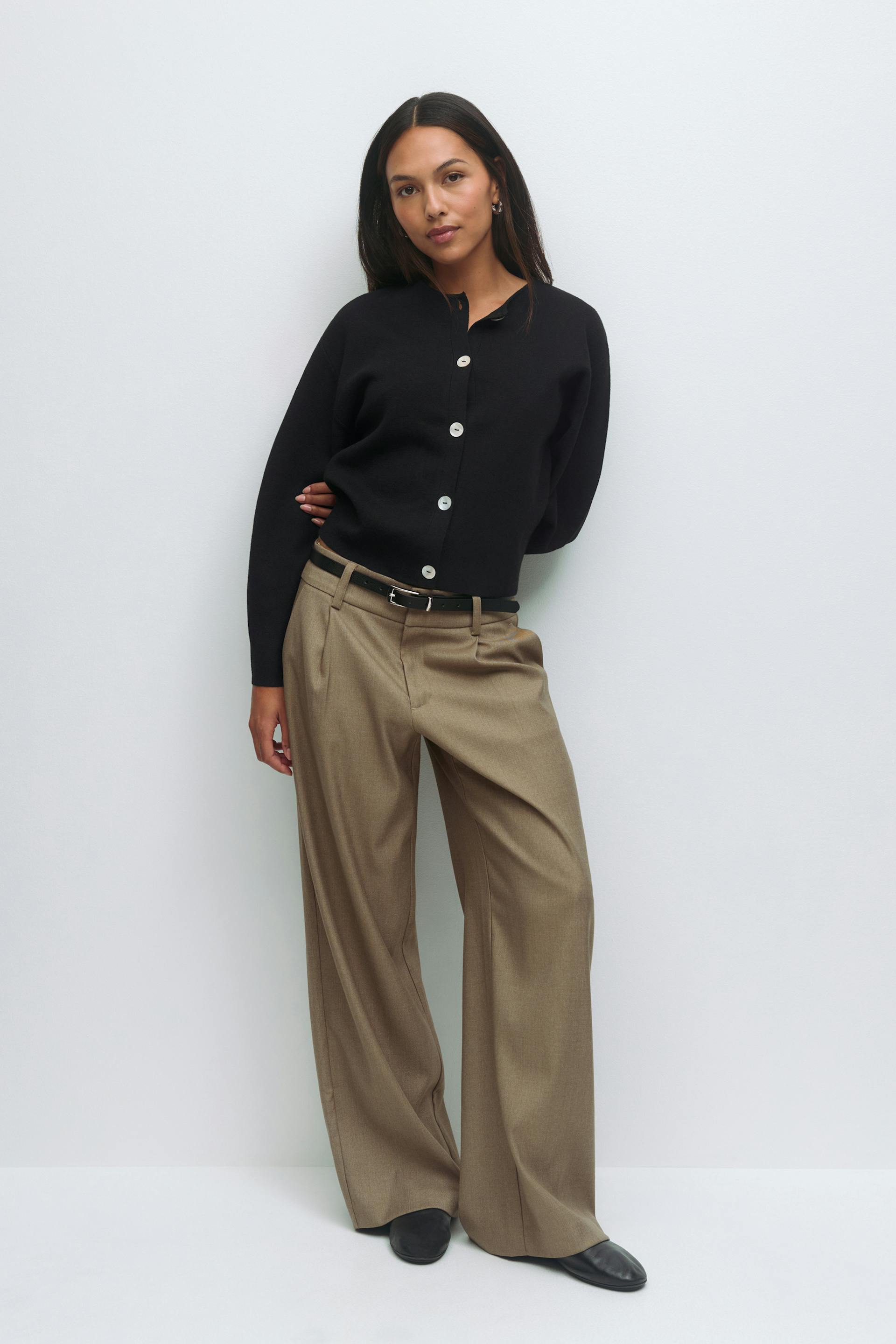 Black long-sleeve buttoned cardigan with white buttons, taupe wide-leg tailored trousers with a black belt, and black pointed-toe flats.