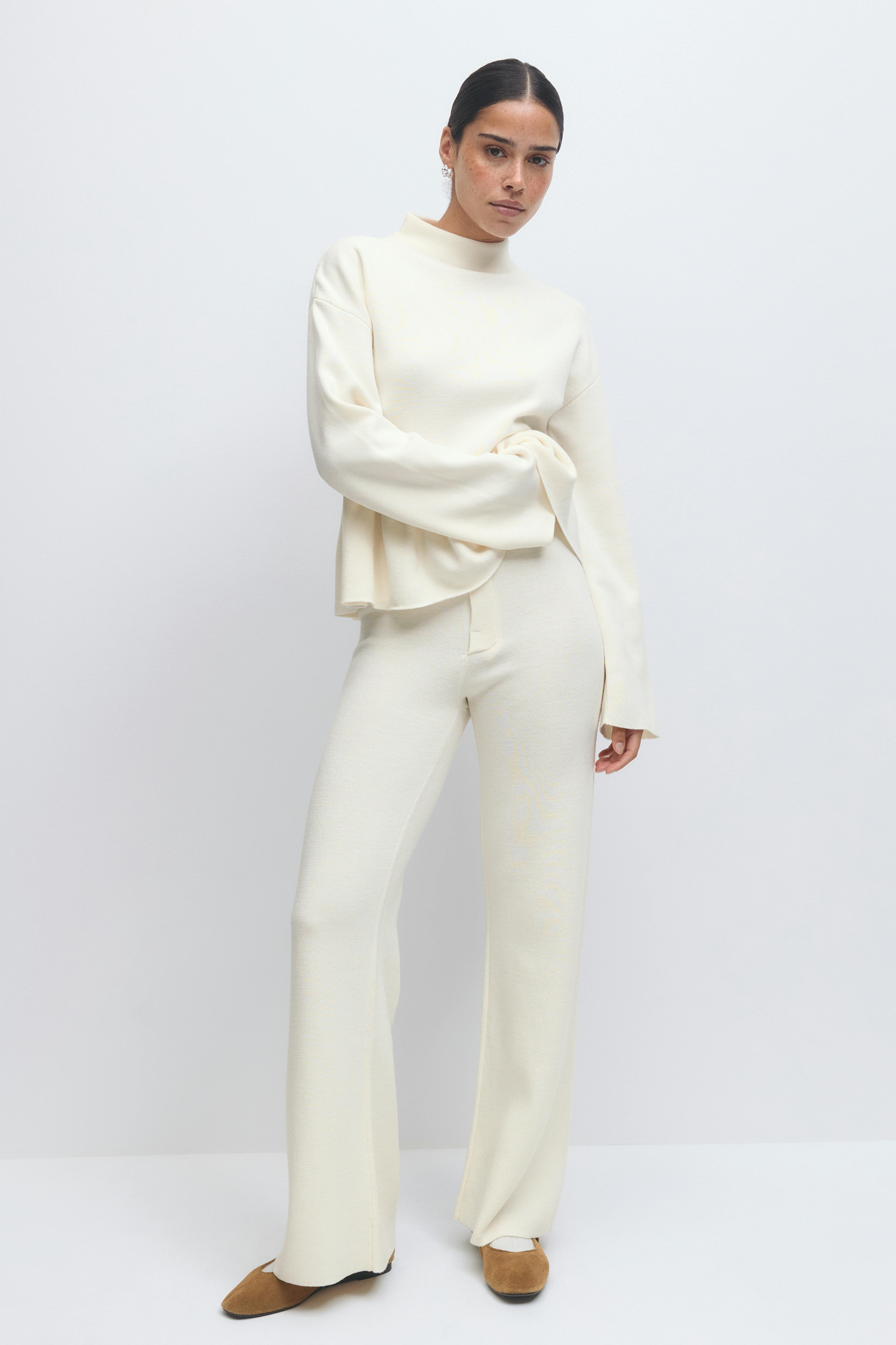 Cremefarbener langärmeliger Pullover mit Mokkakragen, kombiniert mit einer cremefarbenen, weit geschnittenen Strickhose und hellbraunen, spitz zulaufenden Wildlederschuhen.