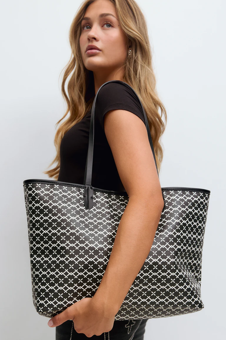 Schwarzes, tailliertes, kurzärmeliges Oberteil, dunkle Denim-Jeans und eine große, schwarze Tote Bag mit weißem, geometrischem Kreuzmuster und schmalen, schwarzen Schulterriemen.