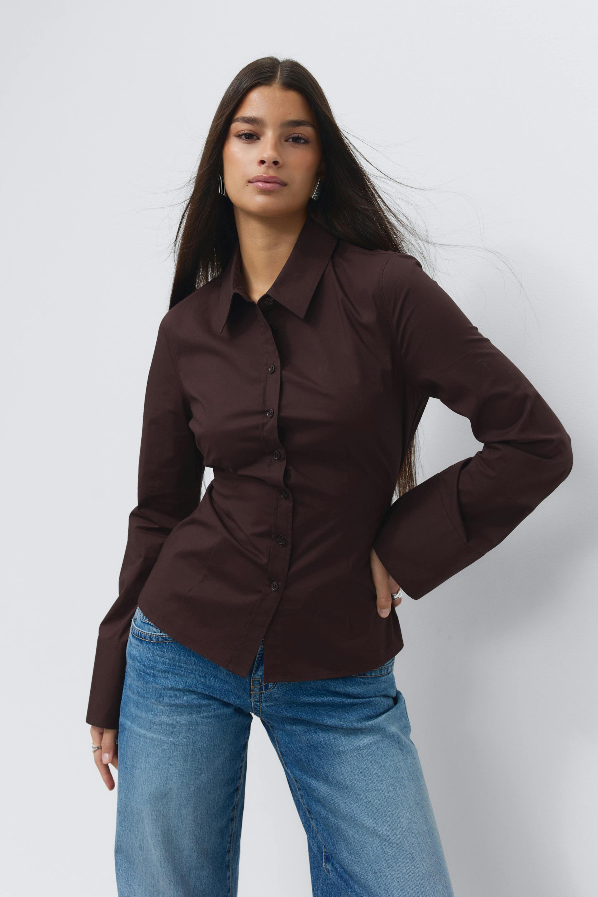 Het model draagt een getailleerd donkerbruin overhemd met lange mouwen en een puntige kraag, gecombineerd met een blauwe jeans met rechte pijpen in medium-wash.