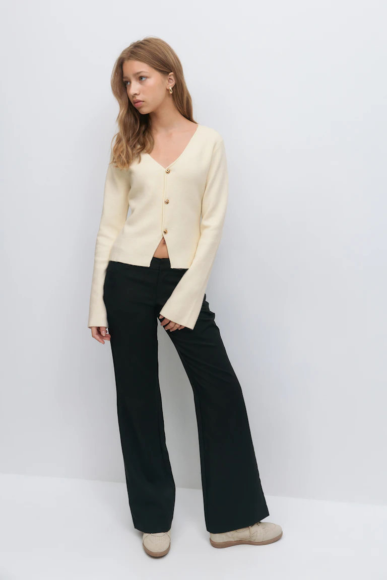 Cremefarbene Strickjacke mit V-Ausschnitt, goldfarbenen Knöpfen und langen Ärmeln, kombiniert mit einer schwarzen Hose mit weitem Bein, beigen Brogue-Schuhen aus Wildleder und goldenen Ohrringen.