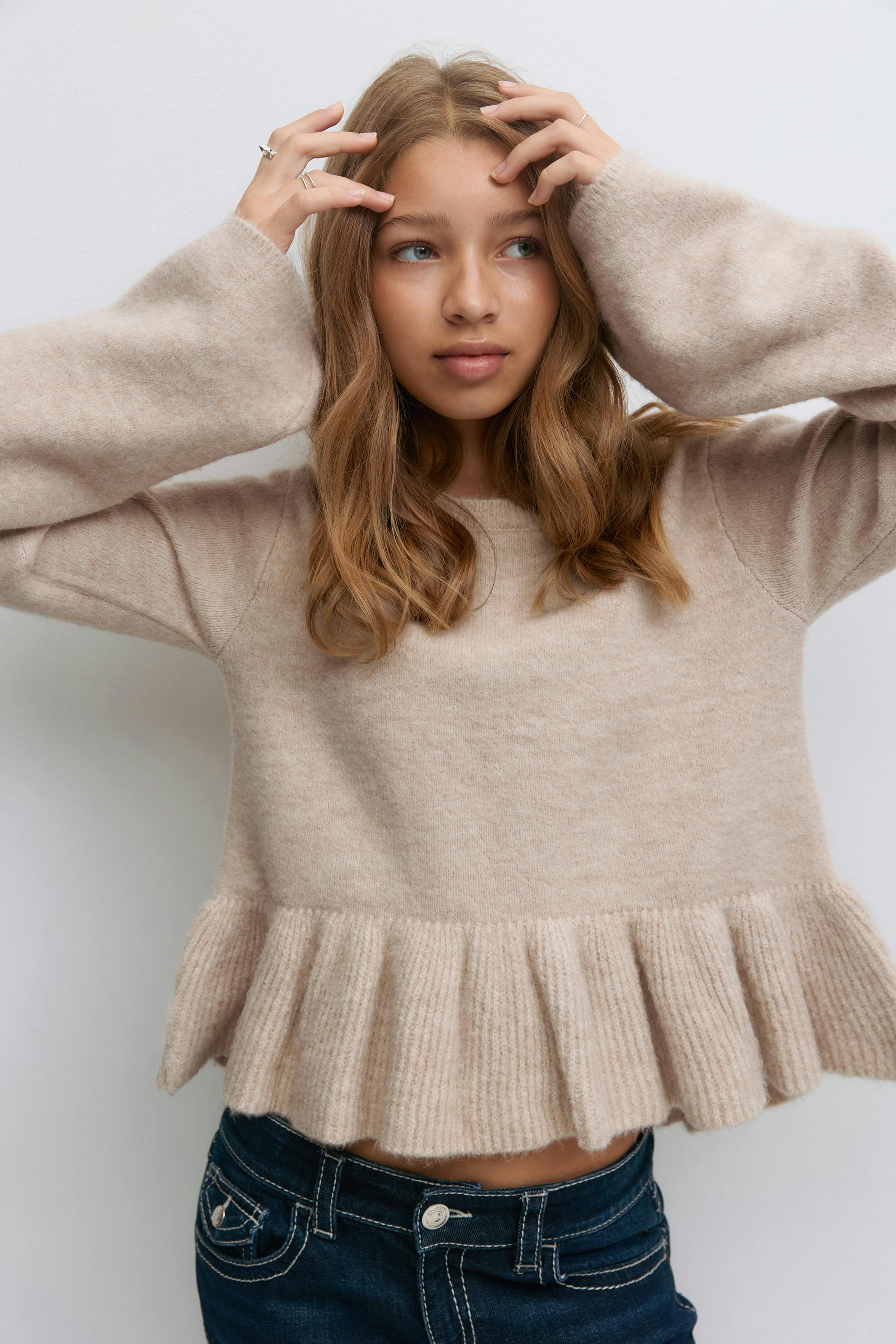 Lys beige fuzzy-strikket sweater med flæsekant og lange udsatte ærmer, sat sammen med mørkeblå denimjeans med kontrastsyninger og knaplukning.