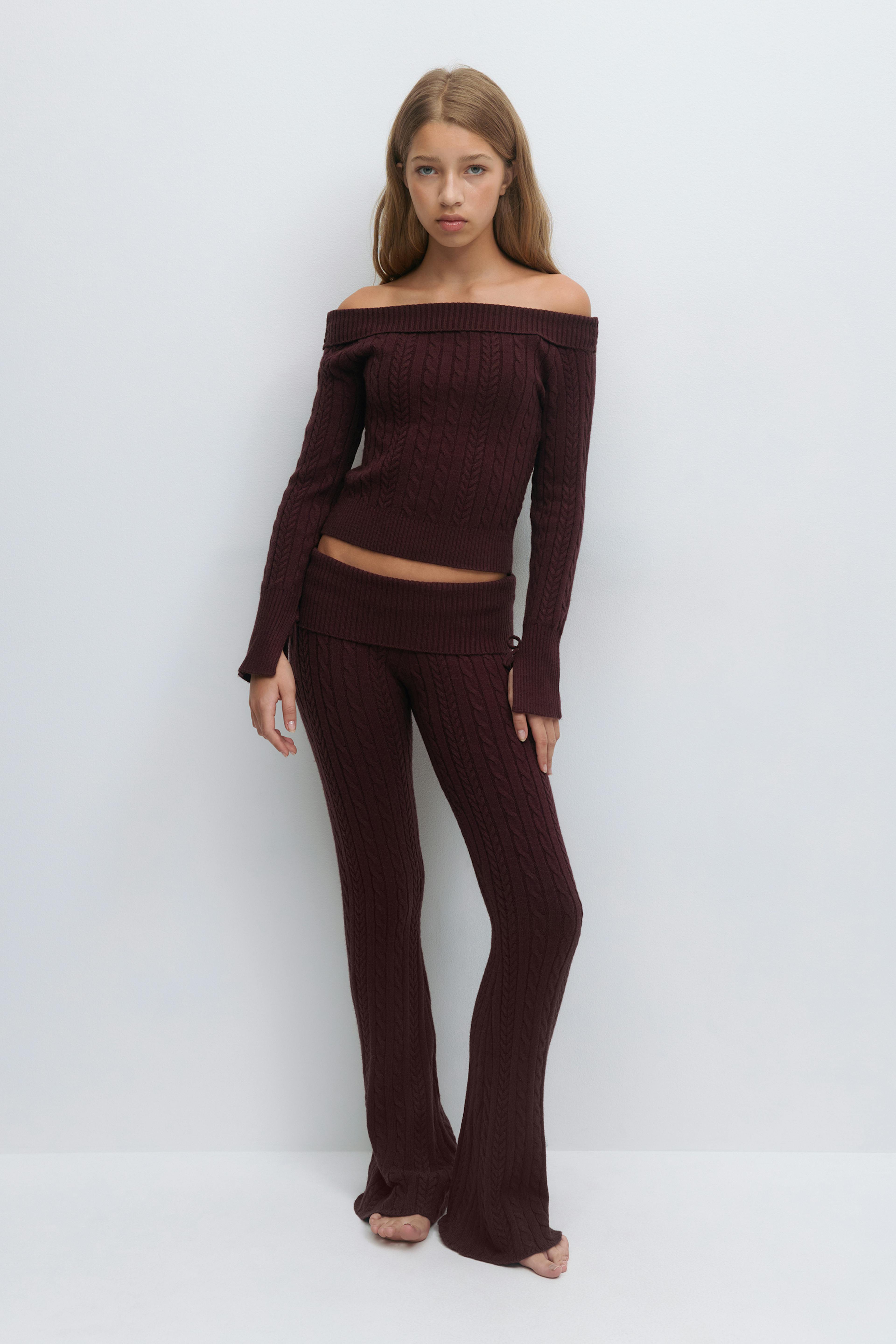 Olkapäätön syvän viininpunainen kaapelineulepusero, jossa on pitkät hihat ja uurrettu helma, yhdistettynä yhteensopiviin korkeavyötäröisiin kaapelineuloksesta leveneviin housuihin.