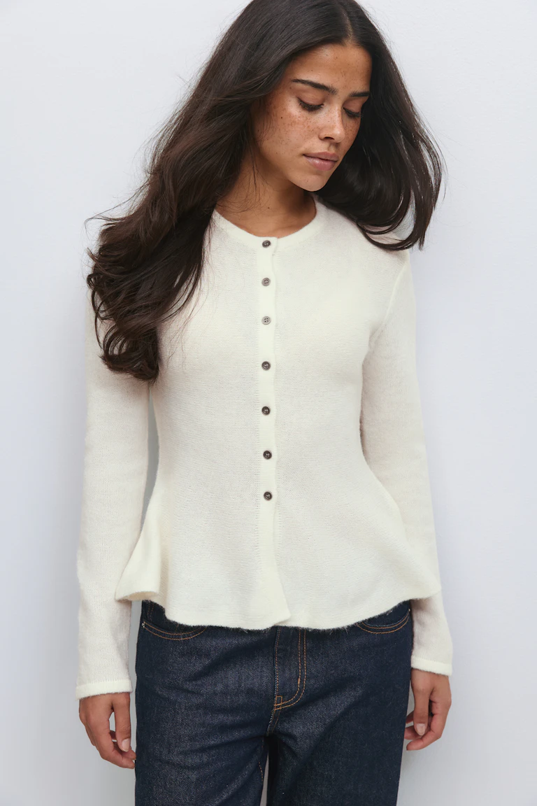 Ivoorkleurig gebreid vest met lange mouwen en knoopsluiting, ronde hals en subtiele peplum zoom, gecombineerd met een donkere indigo spijkerbroek.