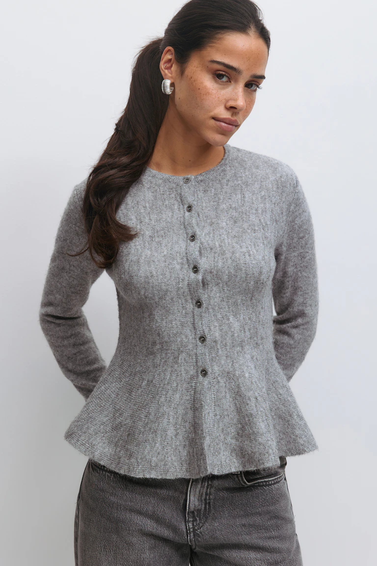 Ljusgrå, långärmad kofta med knappslå, rund halsringning och peplum-fåll, kombinerad med jeans i blekt kol och silverfärgade örhängen med ribbstickning.