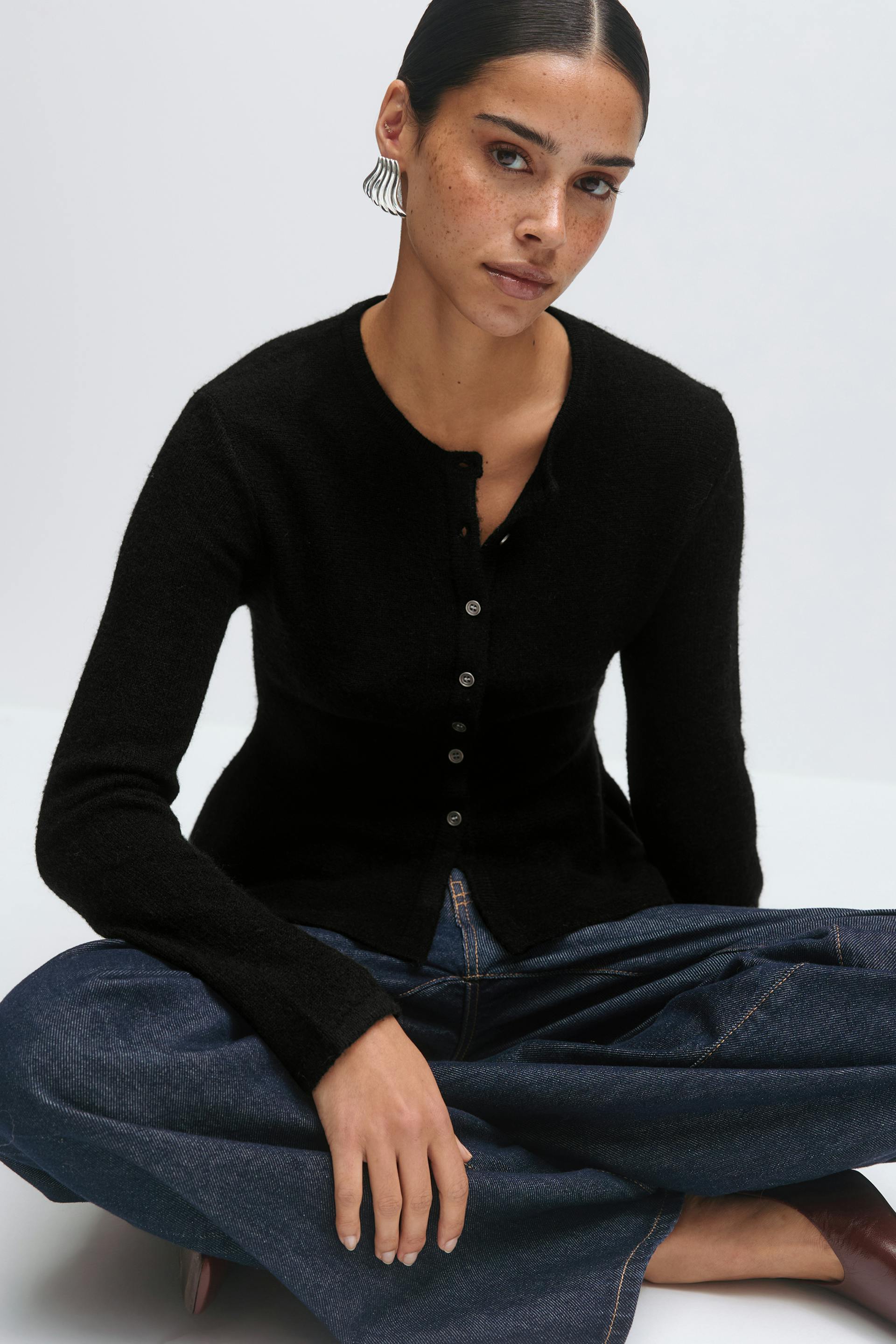 Schwarze, taillierte, langärmelige Strickjacke mit Knopfleiste und Rundhalsausschnitt, gepaart mit dunkelblauen Jeans mit weitem Bein, braunen, spitzen Lederschuhen und silbernen, skulpturalen Ohrringen.