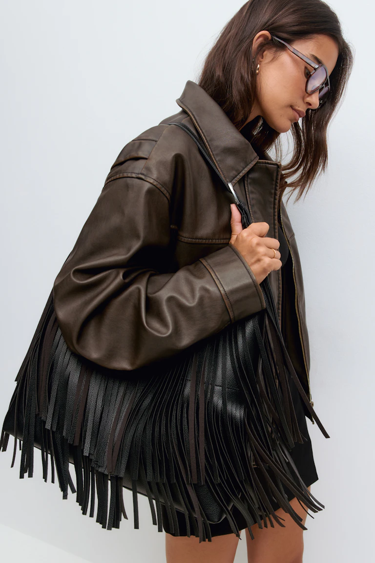 Veste en cuir brun foncé surdimensionnée ; grand sac à bandoulière en cuir noir avec une longue frange ; lunettes de soleil teintées en brun ; mini-jupe noire ; petite boucle d'oreille en or et anneau fin en or.