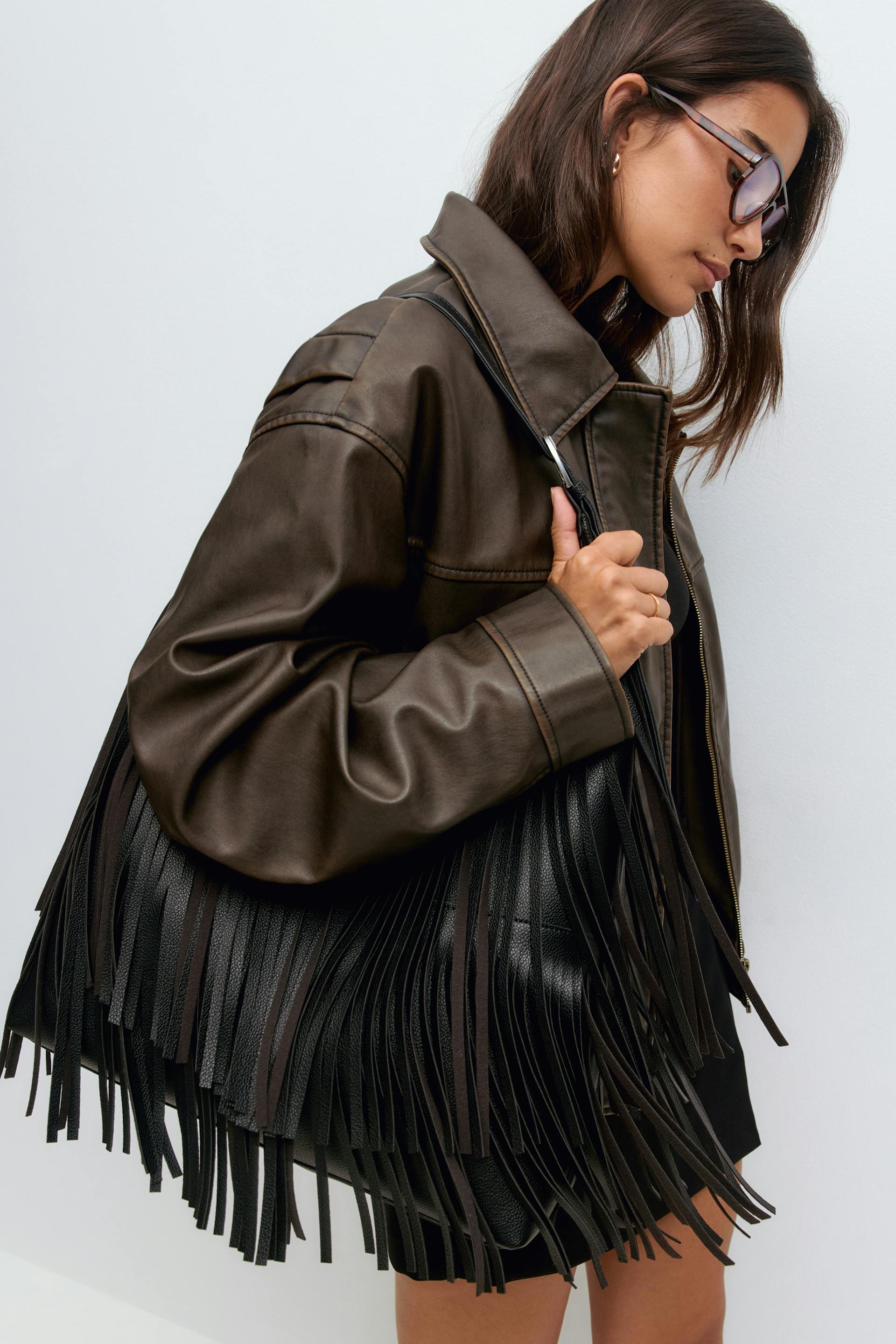 Oversized dark brown leather jacket; large black leather shoulder bag with long fringe; brown-tinted sunglasses; black mini skirt; small gold hoop earring and thin gold band ring.