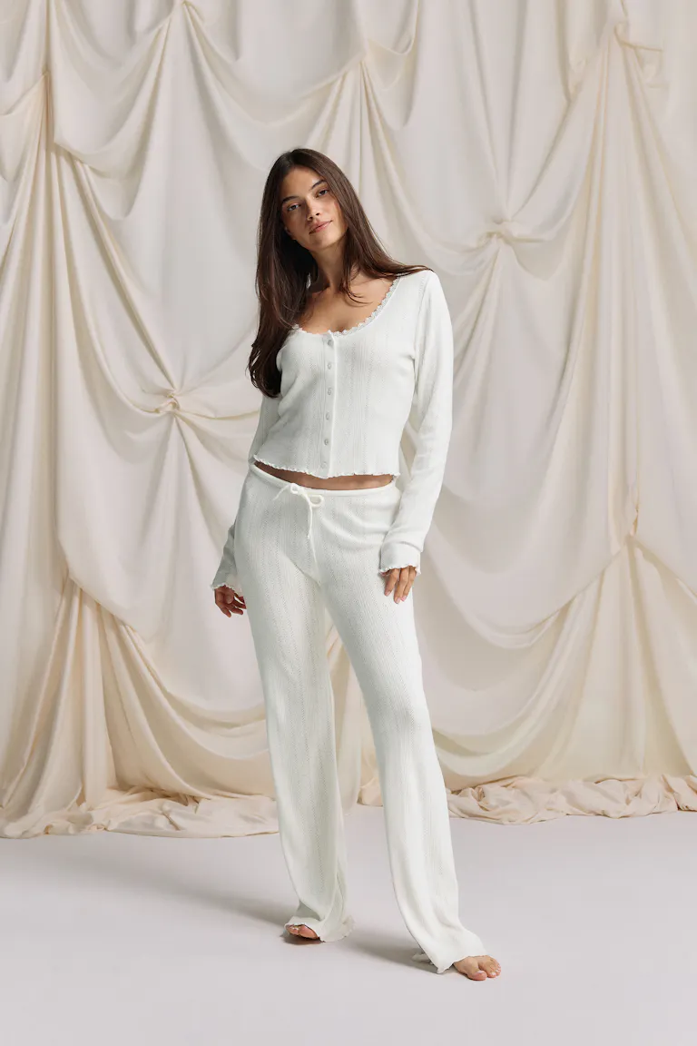 Off-white knit two-piece: a spaghetti-strap camisole with square neckline, front tie and ruffle hem, paired with matching drawstring wide-leg knit trousers with scalloped hems.
