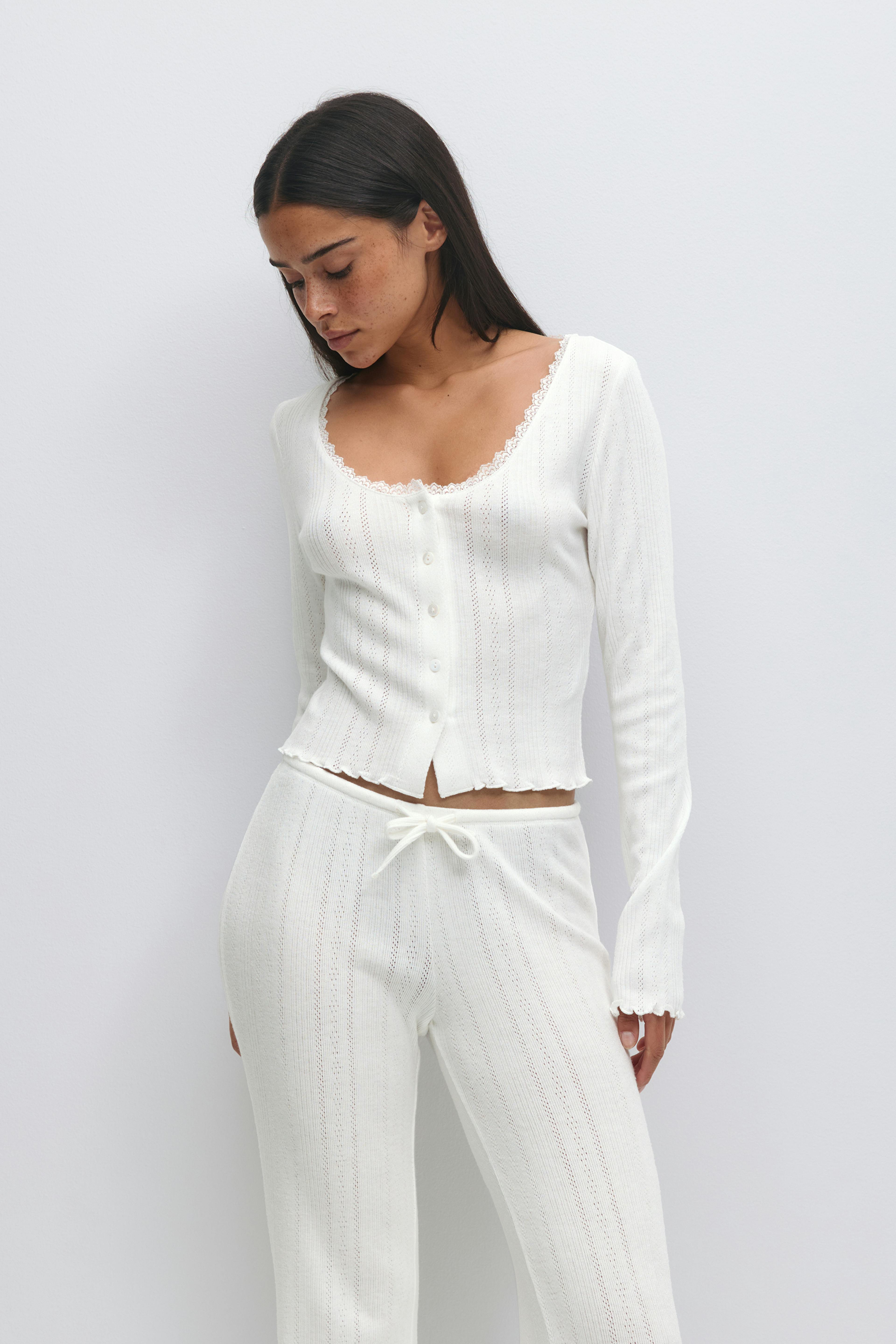 Vit lättviktsstickad tvådelad: långärmad kofta med knappar framtill, skåpsringning och fåll med salladskant, kombinerad med matchande stickade byxor med dragsko i midjan och vertikala öljetter.