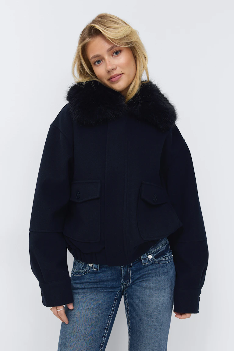 Veste courte en laine marine foncée avec un grand col en fourrure noire, des poches à rabat sur le devant, des manches longues à revers et un ourlet élastiqué, à porter avec un jean bleu délavé.