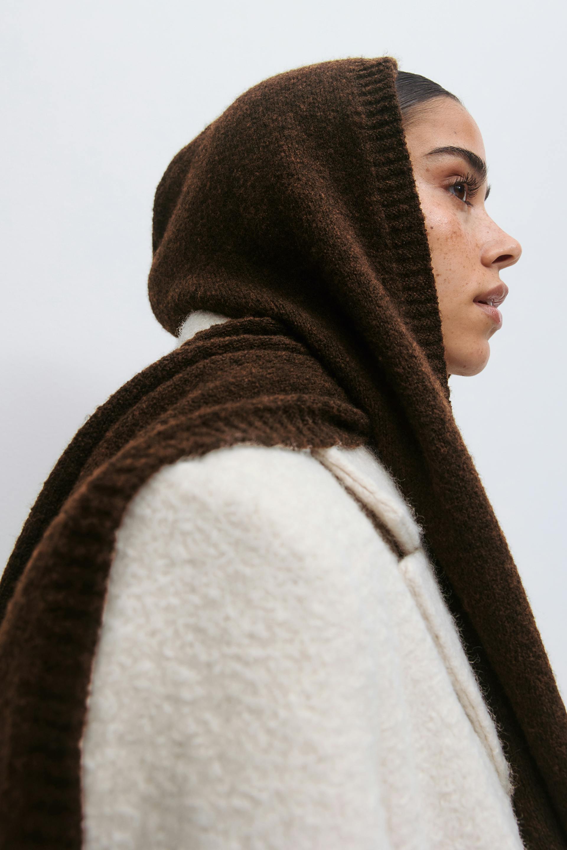 Dark brown knitted hooded scarf draped over the head and shoulders, paired with a cream textured wool coat.