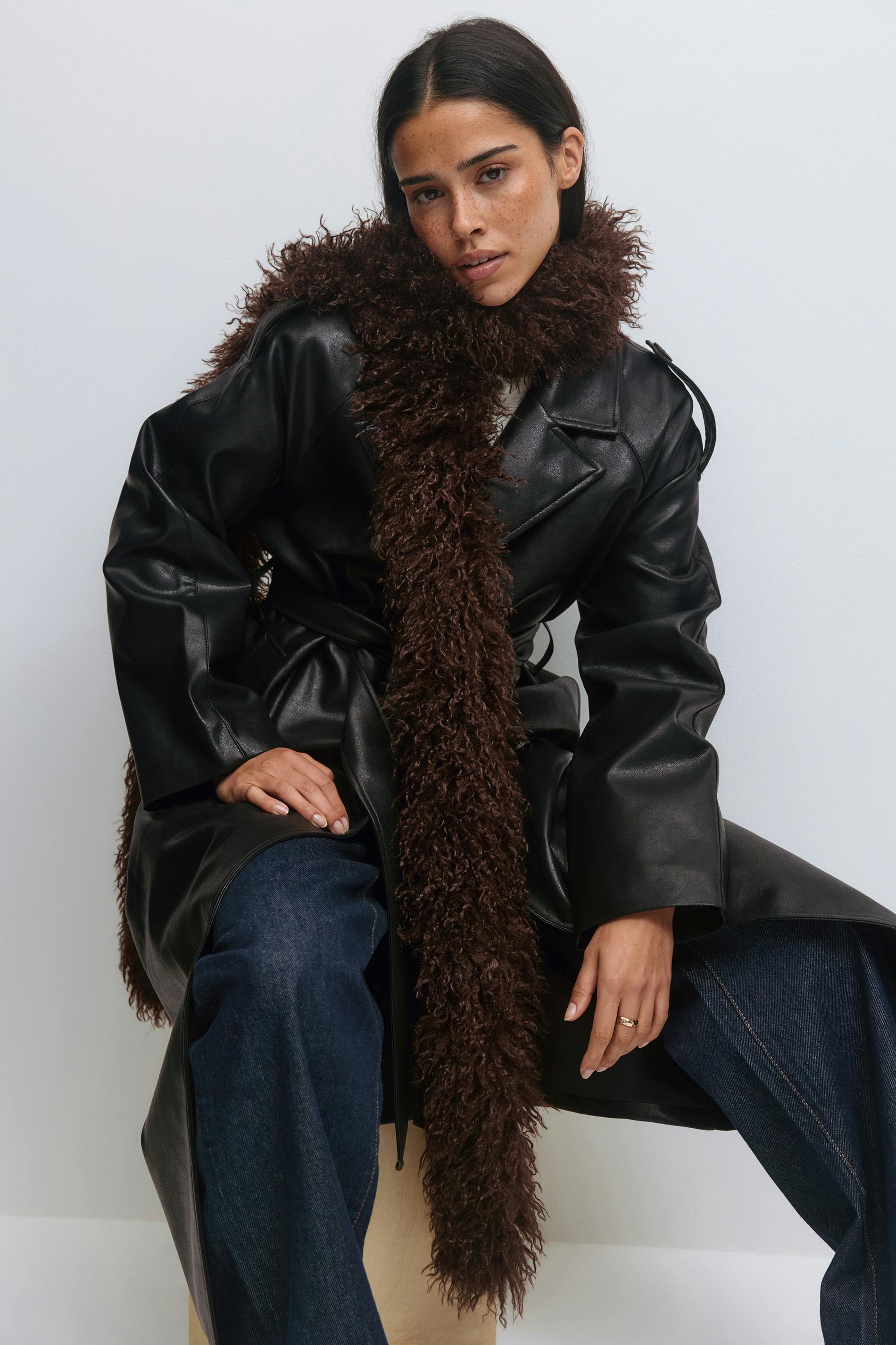 Un trench-coat en cuir noir ceinturé à larges revers, une longue écharpe en fausse fourrure hirsute marron foncé et un jean large bleu foncé.