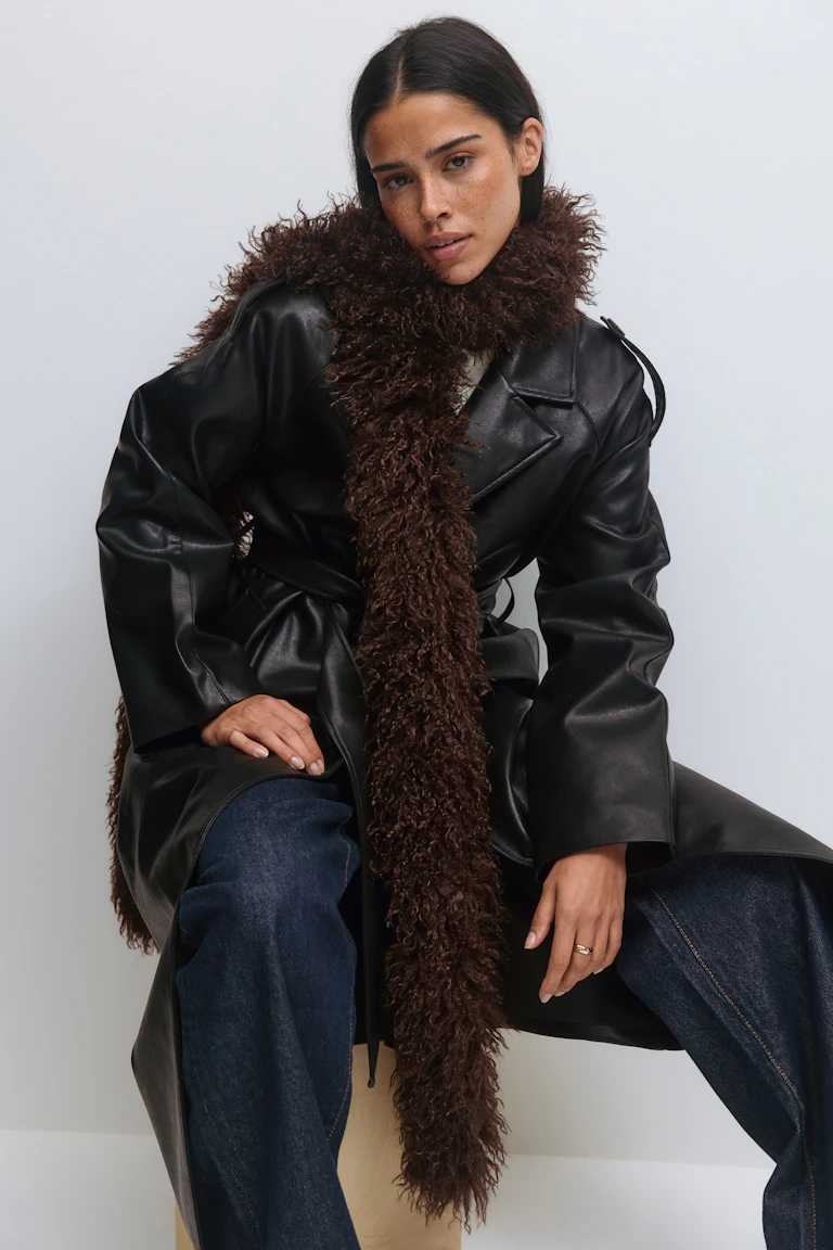 Schwarzer Trenchcoat aus Leder mit Gürtel und breitem Revers, ein langer dunkelbrauner zotteliger Kunstpelzschal und dunkelblaue Jeans mit weitem Bein.